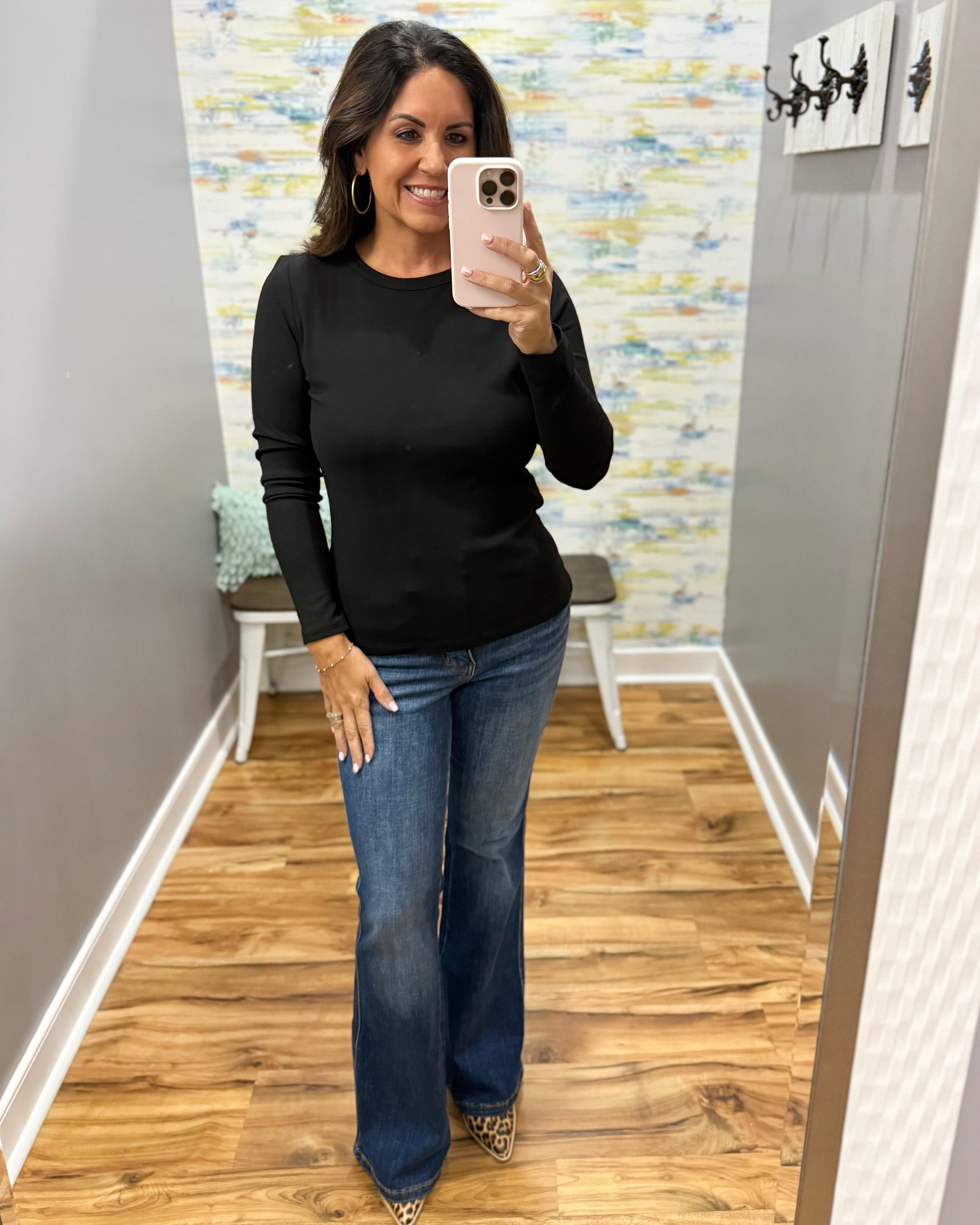 A woman takes a mirror selfie wearing a black long-sleeve crewneck top and blue flare jeans in a fitting room with wood floors and a patterned wall.