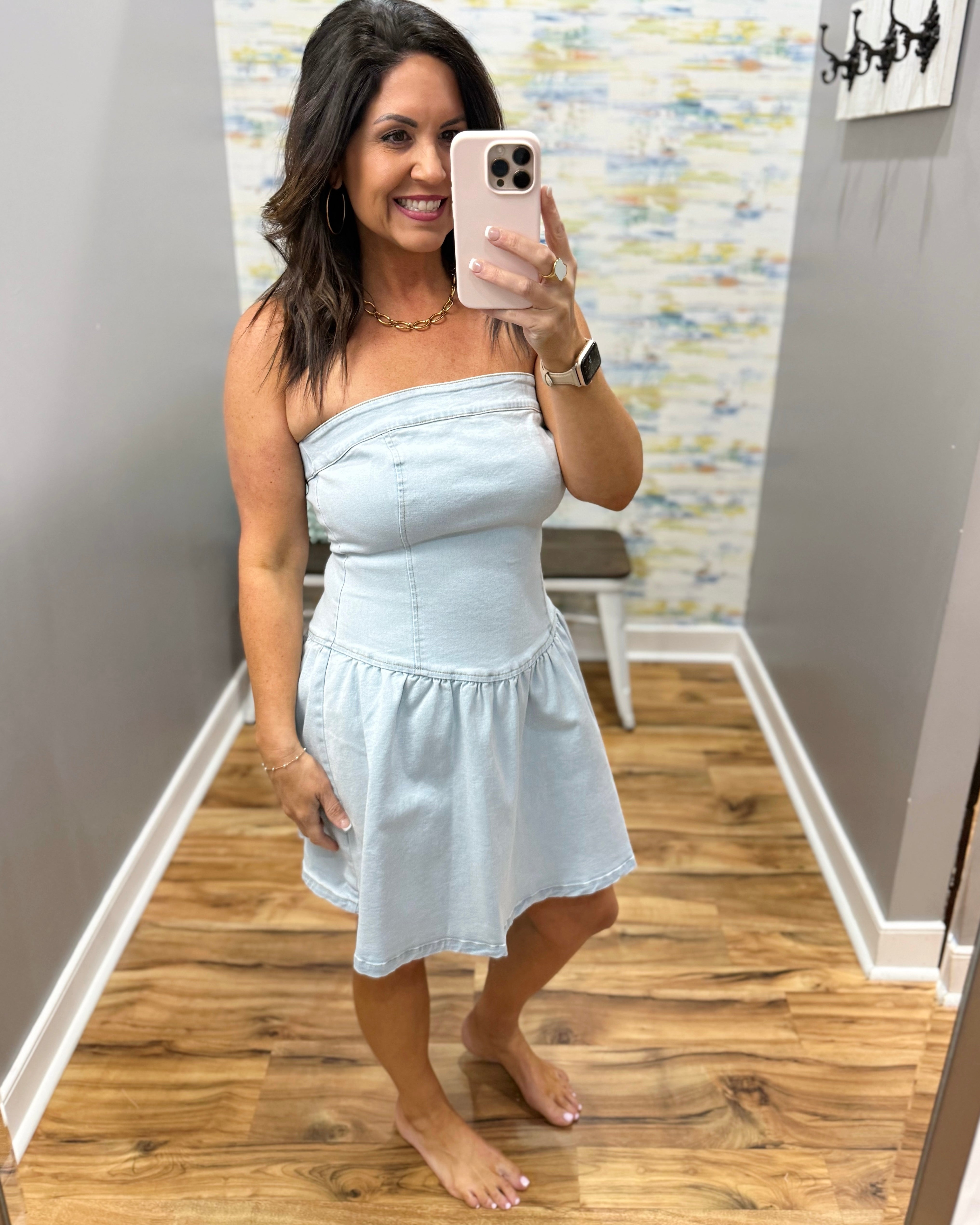 Woman taking a mirror selfie wearing a light blue strapless dress in a room with wooden flooring and a colorful wall.