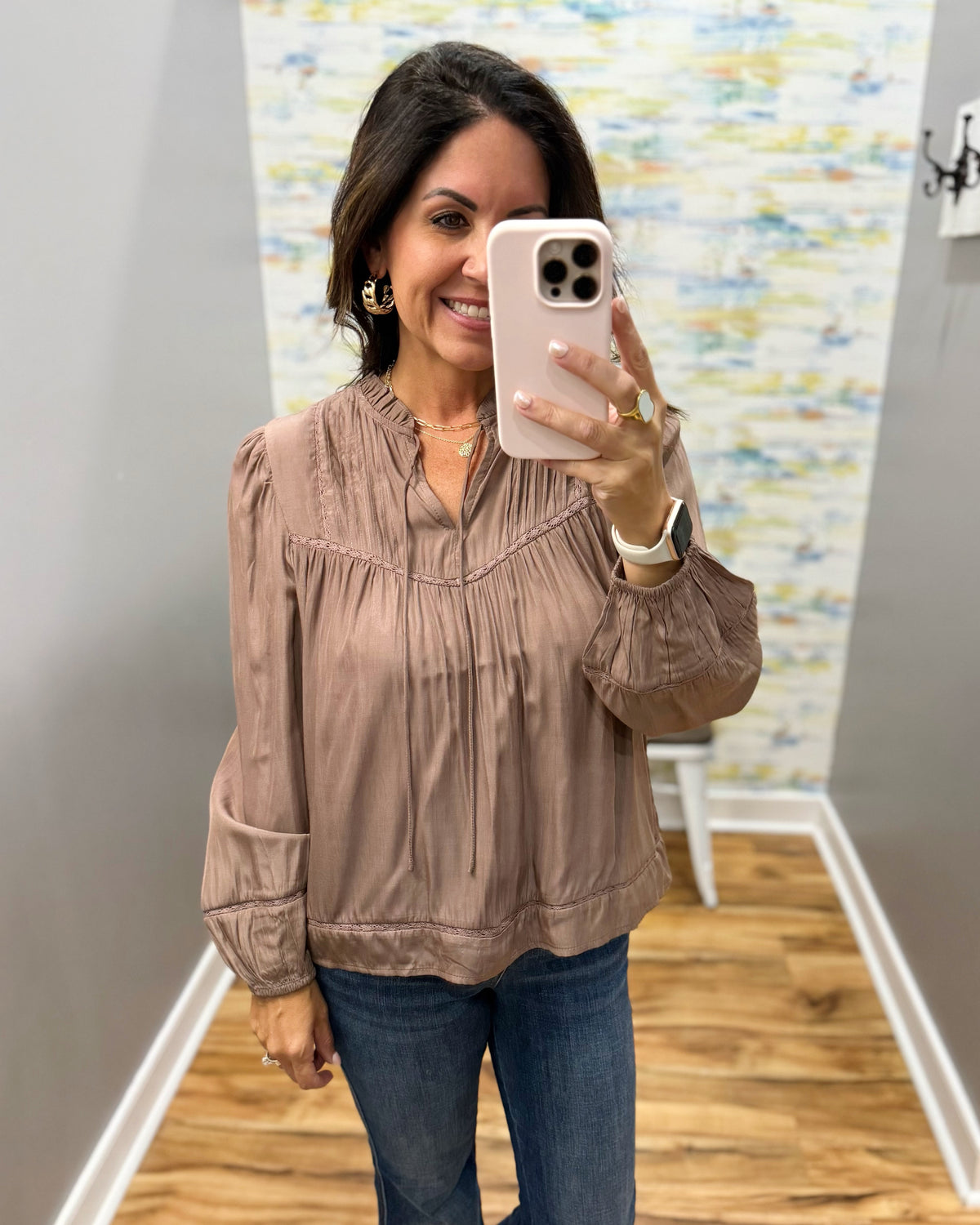 A woman takes a mirror selfie wearing a dusty rose long-sleeve blouse with a gathered, pleated neckline and blue jeans in a home setting with wood floors and gray walls.