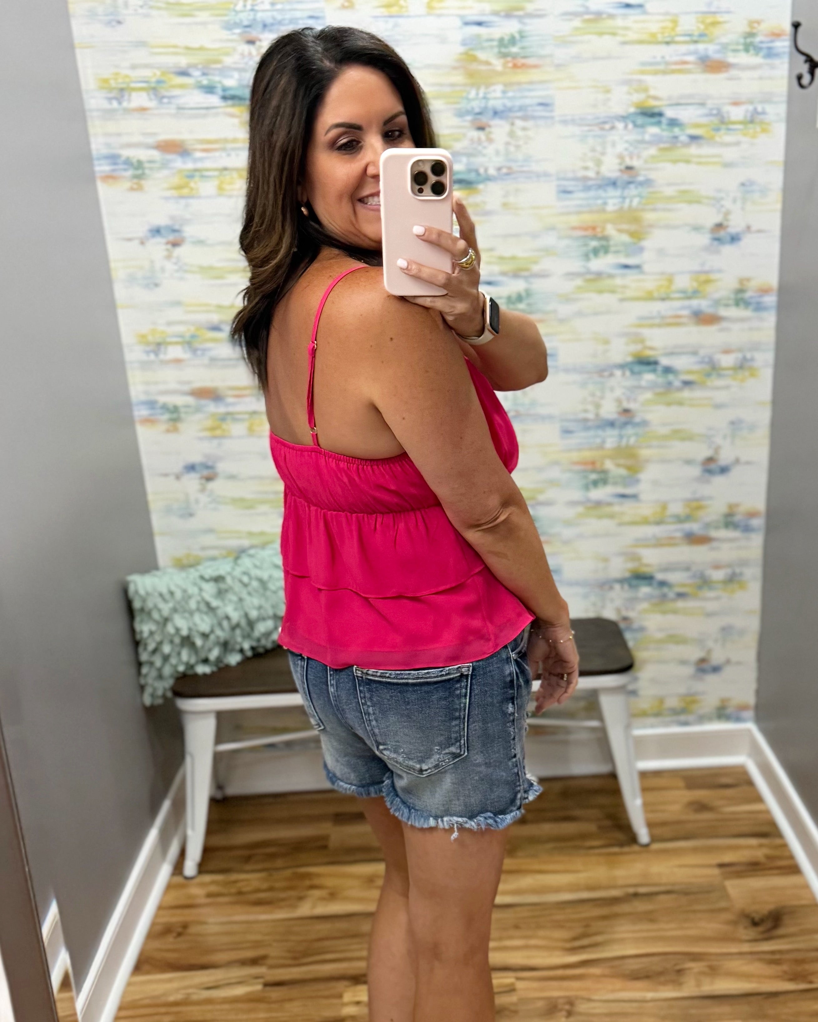 Woman taking a mirror selfie wearing a pink top and denim shorts in a room with a colorful wall.