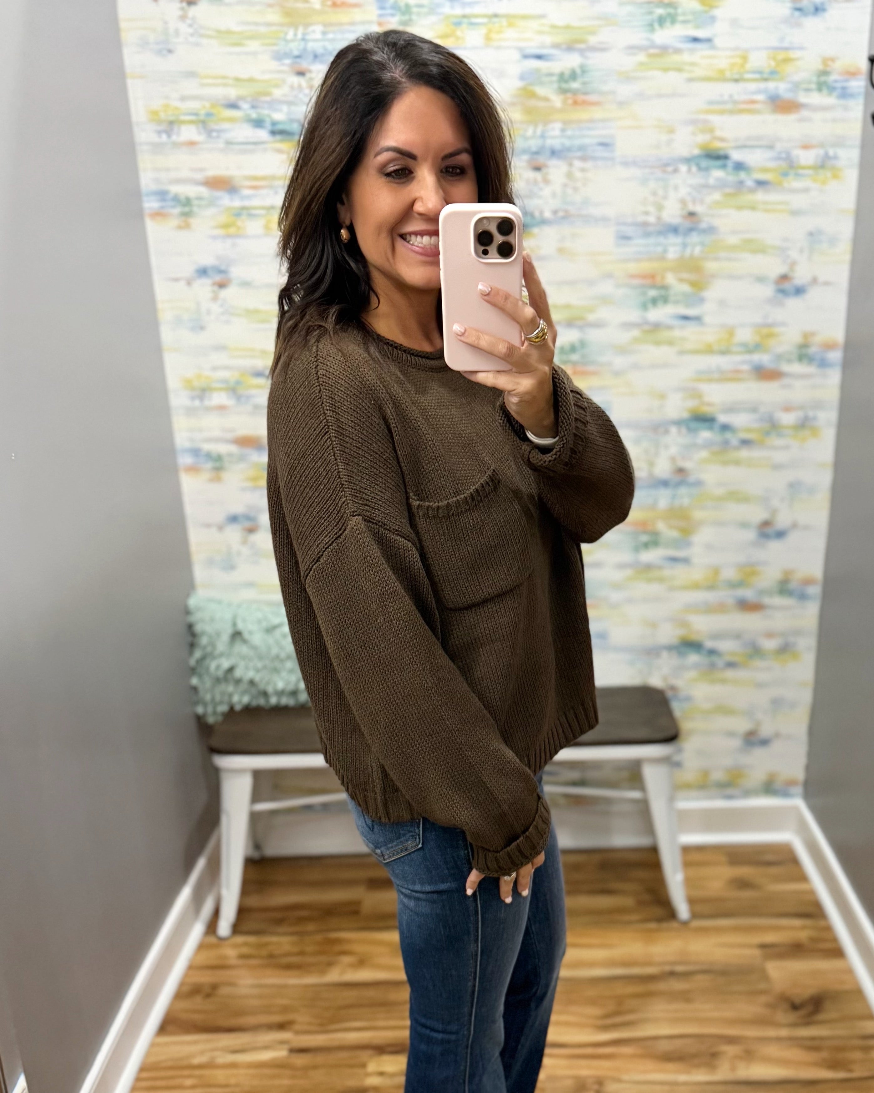 A woman takes a mirror selfie wearing a dark brown knit crewneck sweater and blue jeans in a fitting room with a patterned wallpaper background.