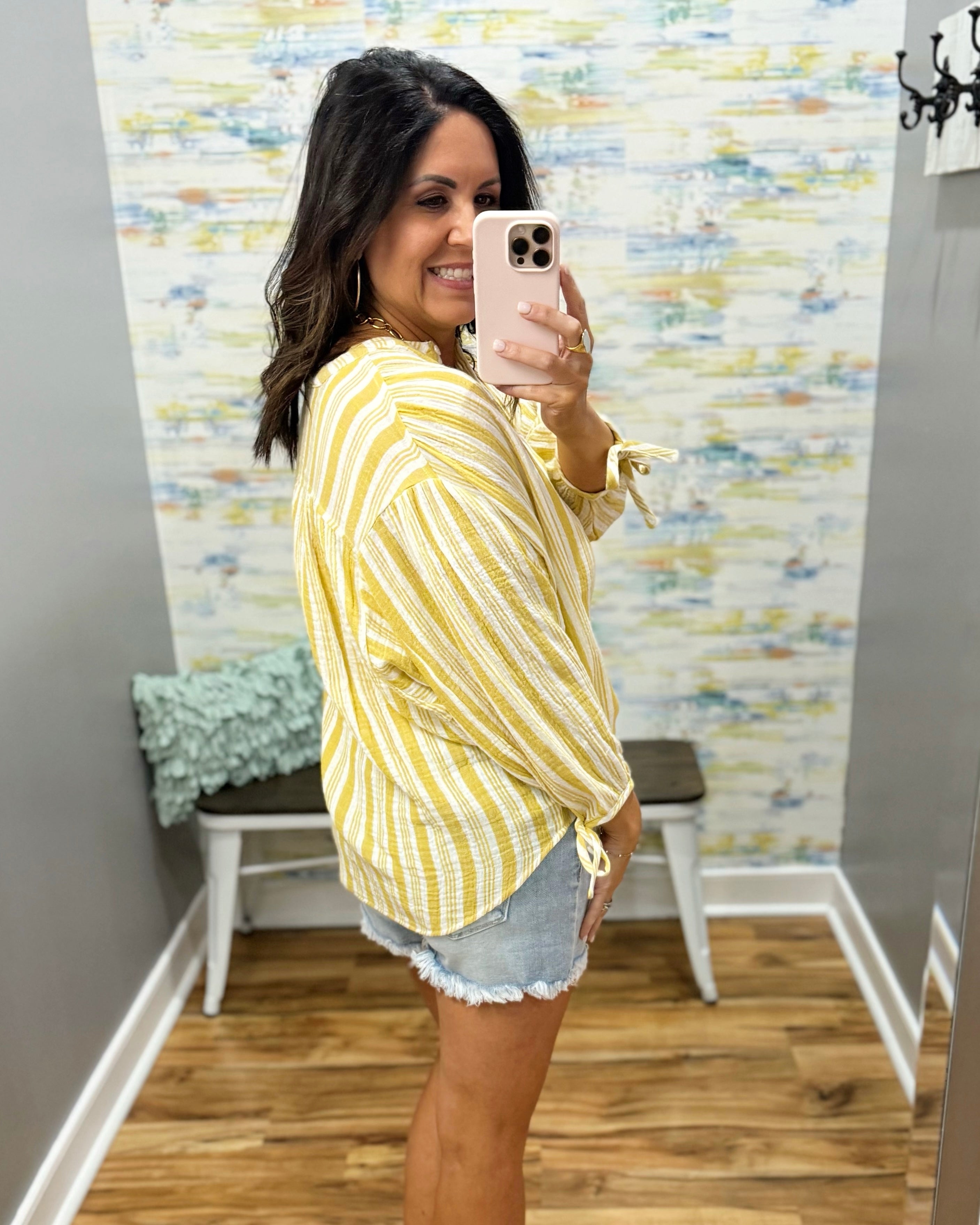 Woman taking a mirror selfie wearing a yellow striped shirt and denim shorts in a room with a textured wall.
