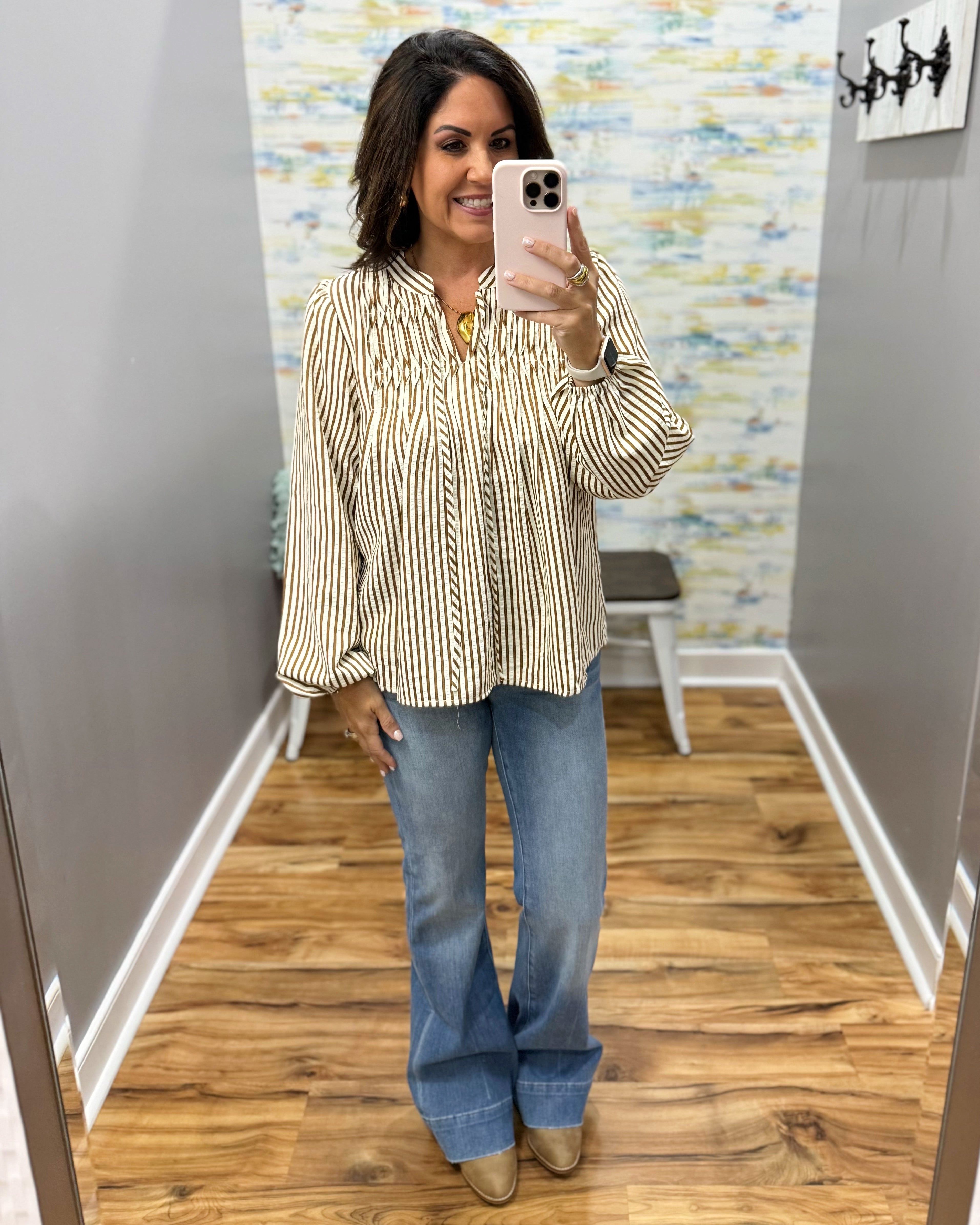 A woman takes a mirror selfie in a fitting room wearing a beige and black vertically striped button-down blouse with balloon sleeves, paired with blue flare jeans and tan boots.