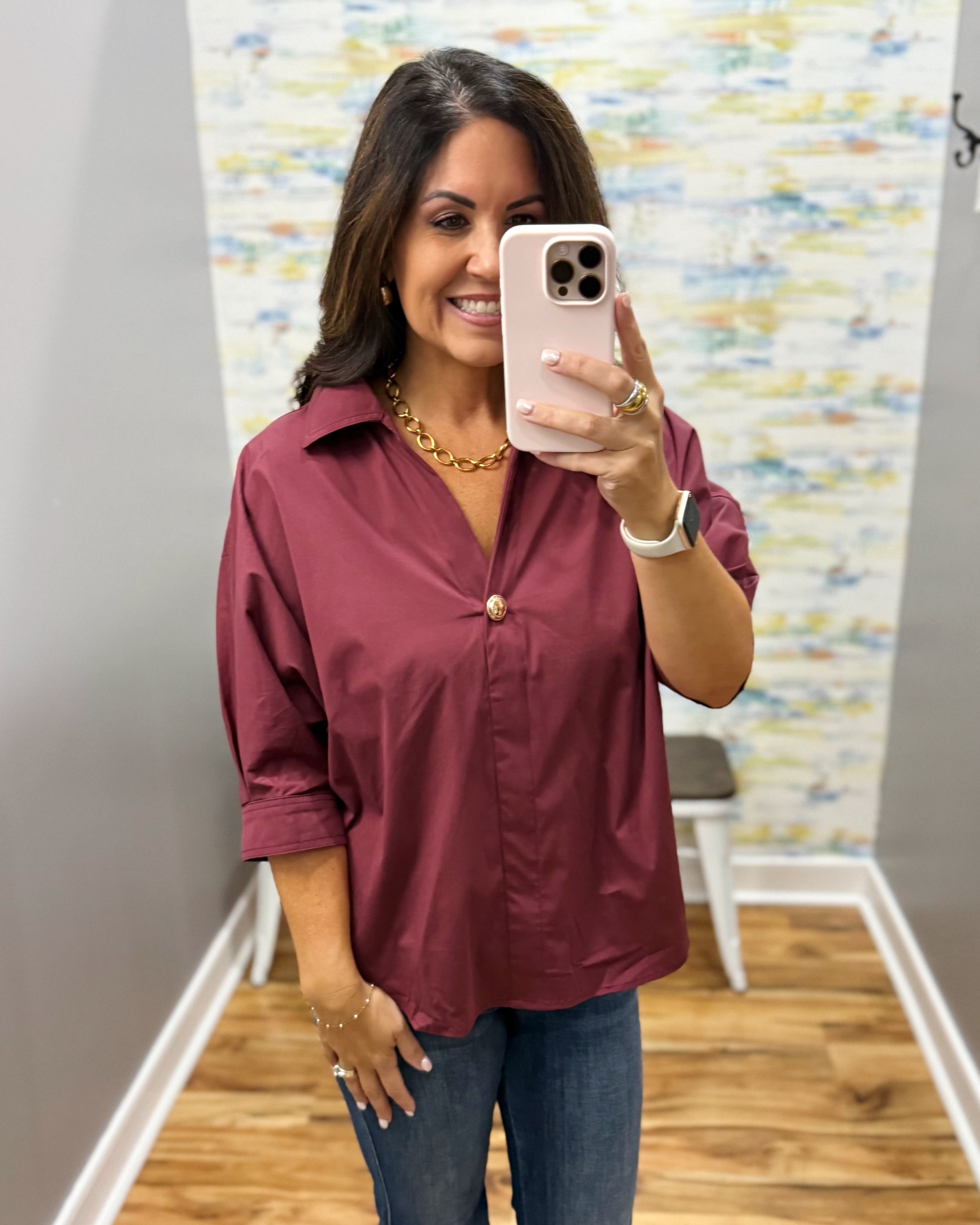 A woman takes a mirror selfie wearing a burgundy button-down shirt with rolled-up sleeves and blue jeans, standing in a fitting room or boutique.