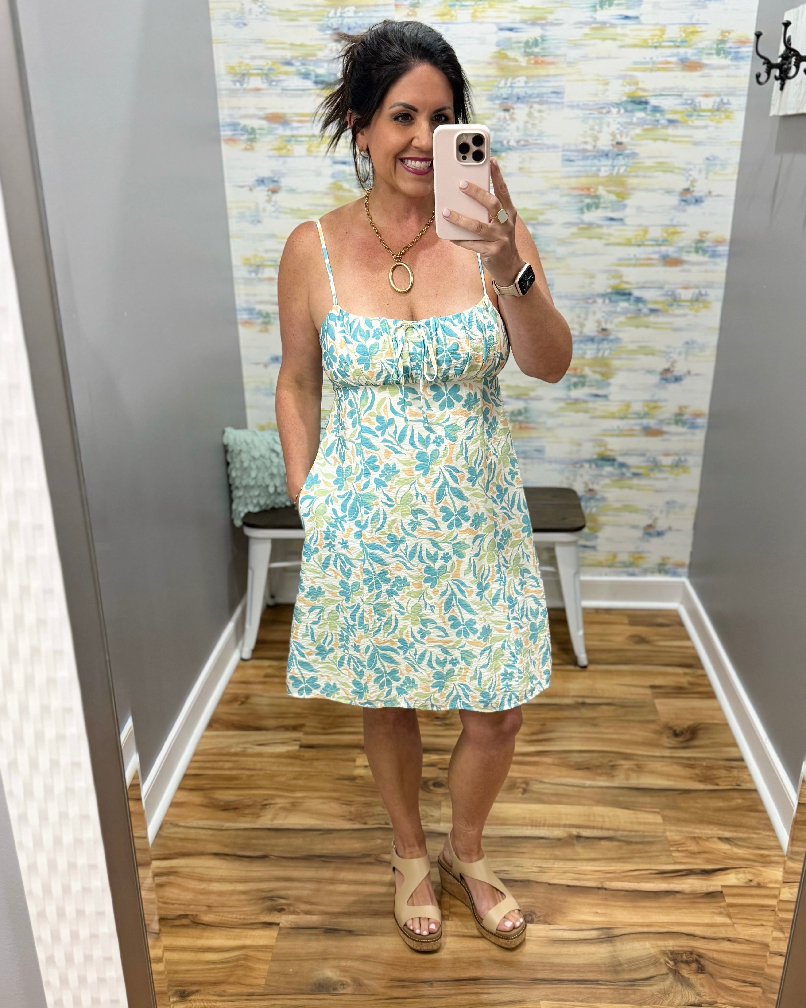 Woman taking a mirror selfie wearing a floral dress in a room with a patterned wall.