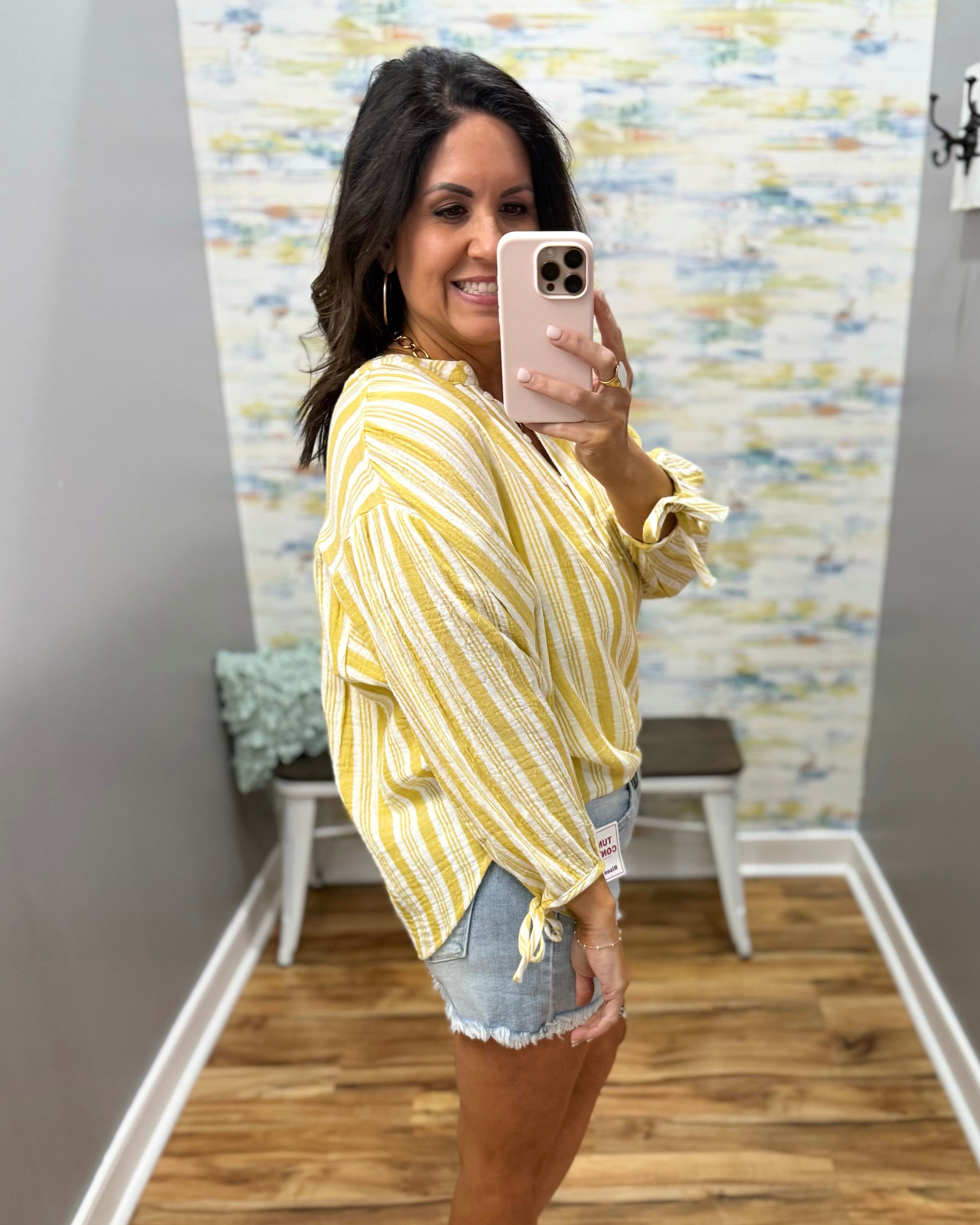 Woman taking a mirror selfie wearing a yellow patterned top and denim shorts in a room with a colorful wall.