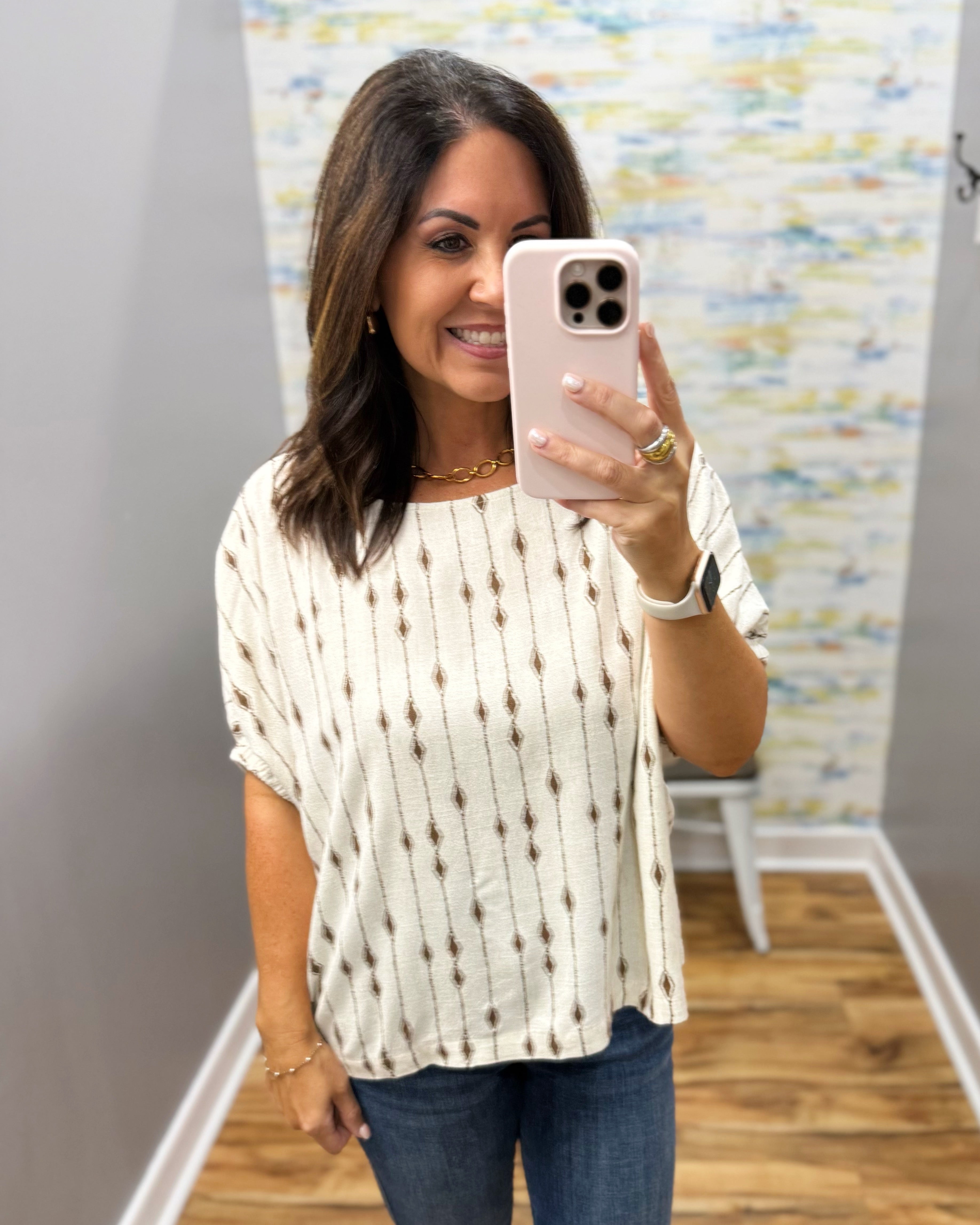 A woman taking a mirror selfie wearing a cream-colored short-sleeve top with a brown vertical dotted pattern, paired with blue jeans.