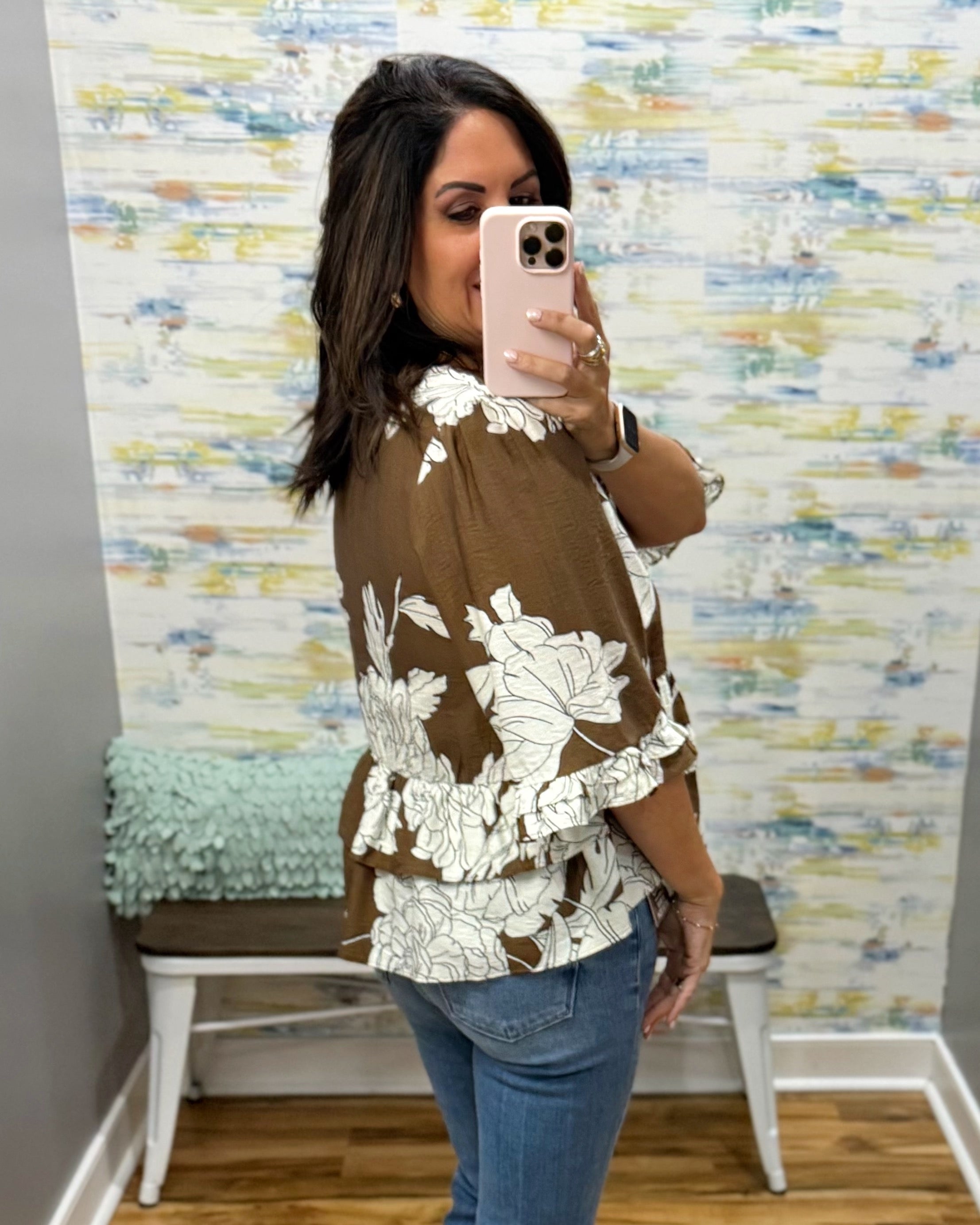 A woman takes a mirror selfie in a boutique showing a brown and white floral print blouse, paired with blue jeans, in front of a patterned wallpaper background.