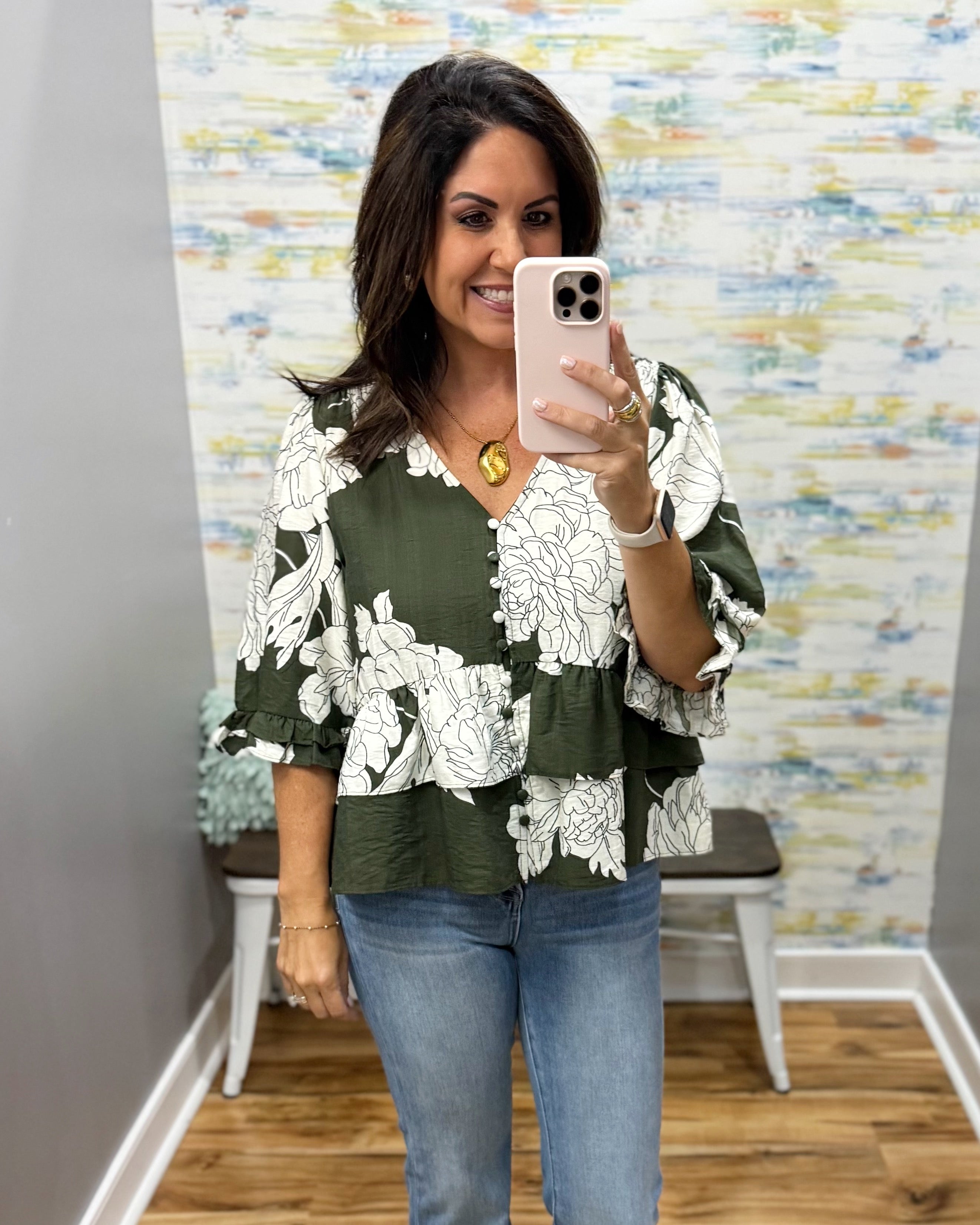 A woman takes a mirror selfie wearing a green and white floral print blouse with ruffled sleeves and blue jeans, standing in front of a patterned wall.