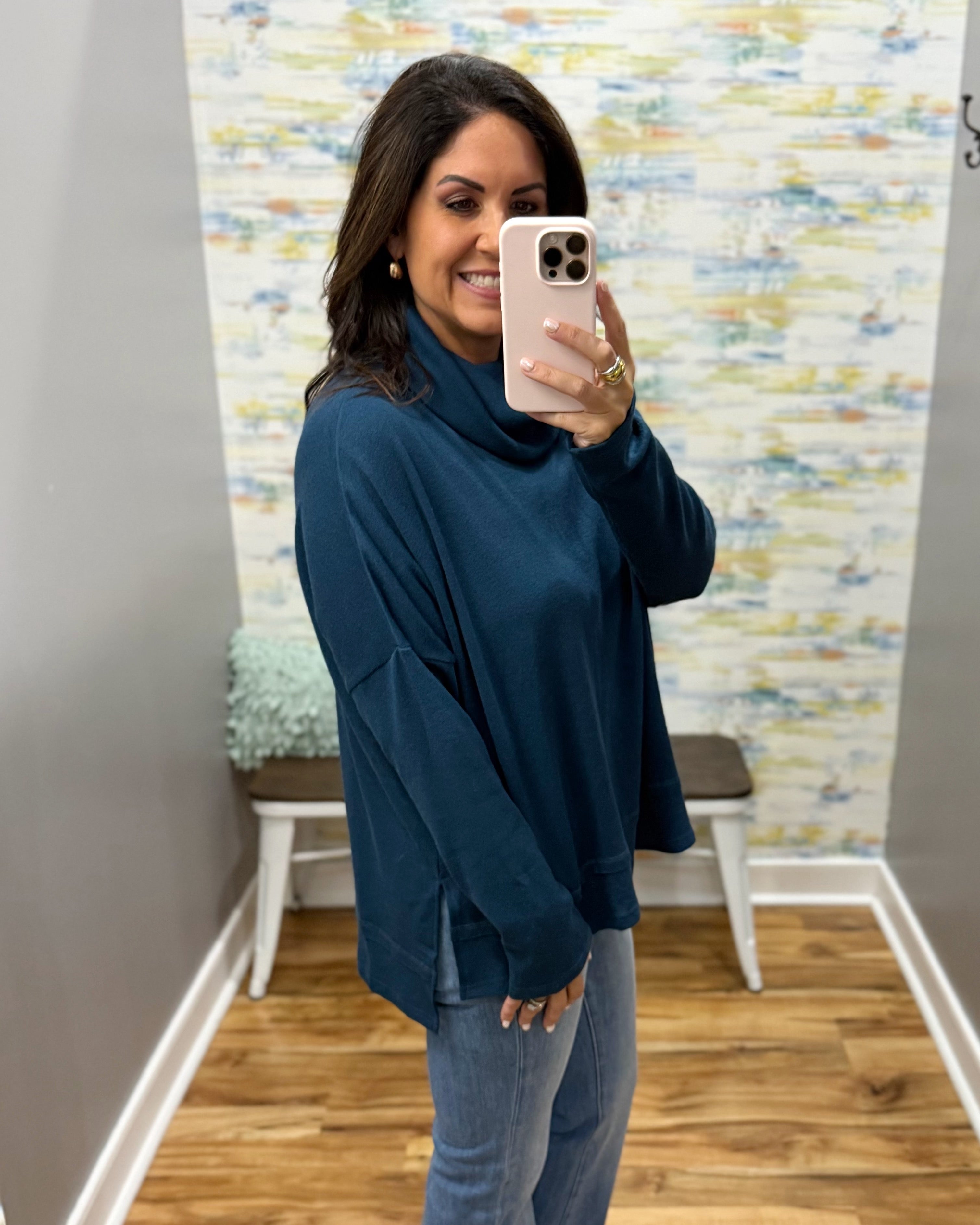 A woman takes a mirror selfie wearing a dark teal long-sleeve turtleneck top and light blue jeans in a fitting room with wood floors and a patterned wallpaper background.