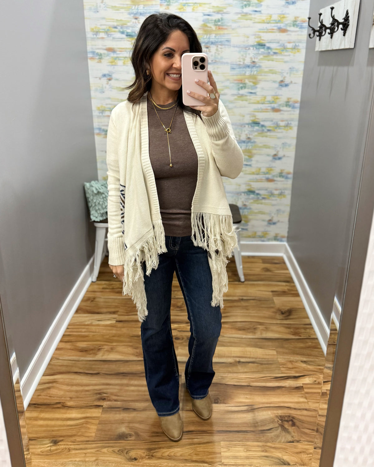 A woman takes a mirror selfie in a fitting room wearing a cream knit cardigan with fringe, a brown ribbed top, dark blue jeans, and tan ankle boots.