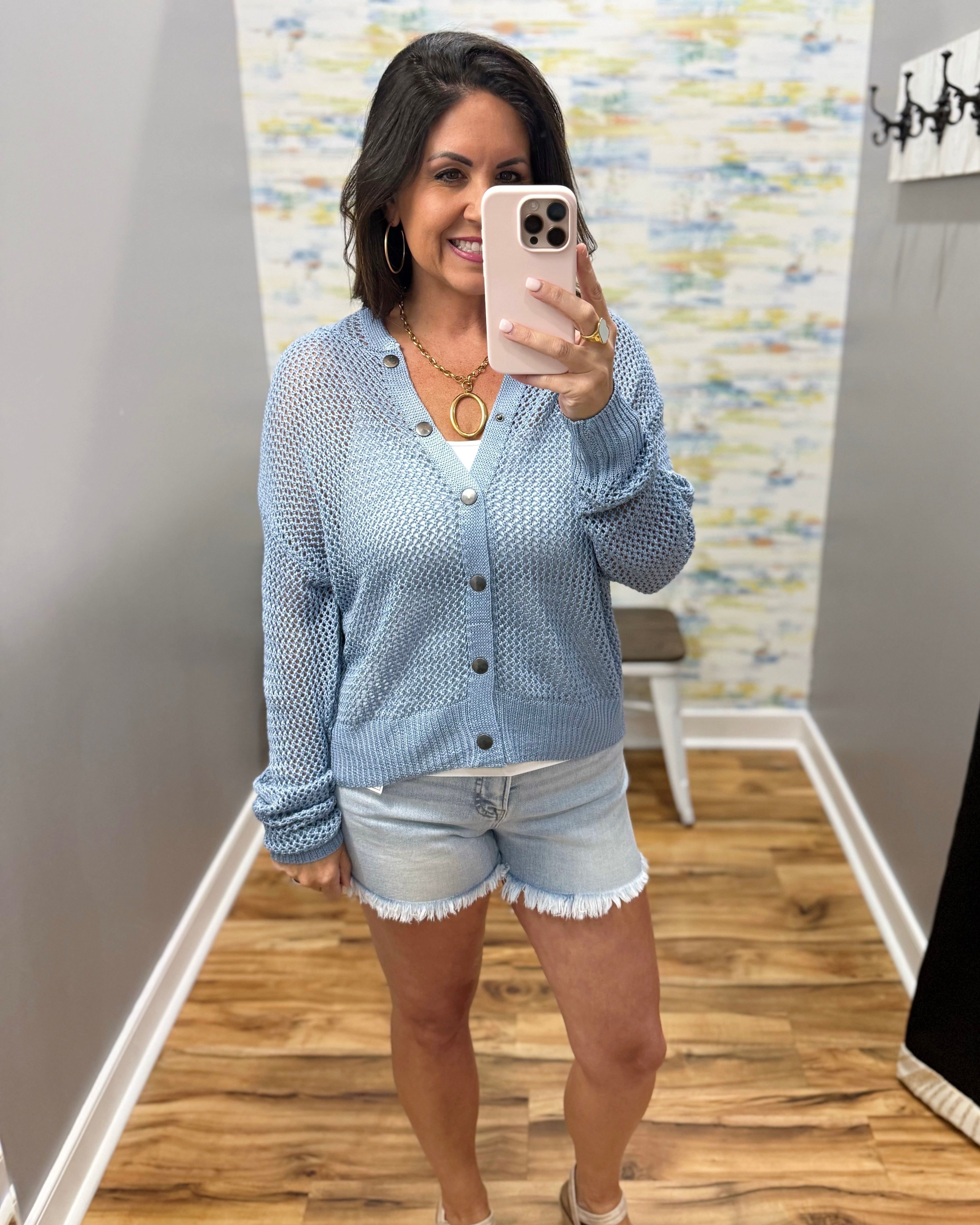 Woman taking a mirror selfie wearing a light blue cardigan and denim shorts in a room with patterned wallpaper.