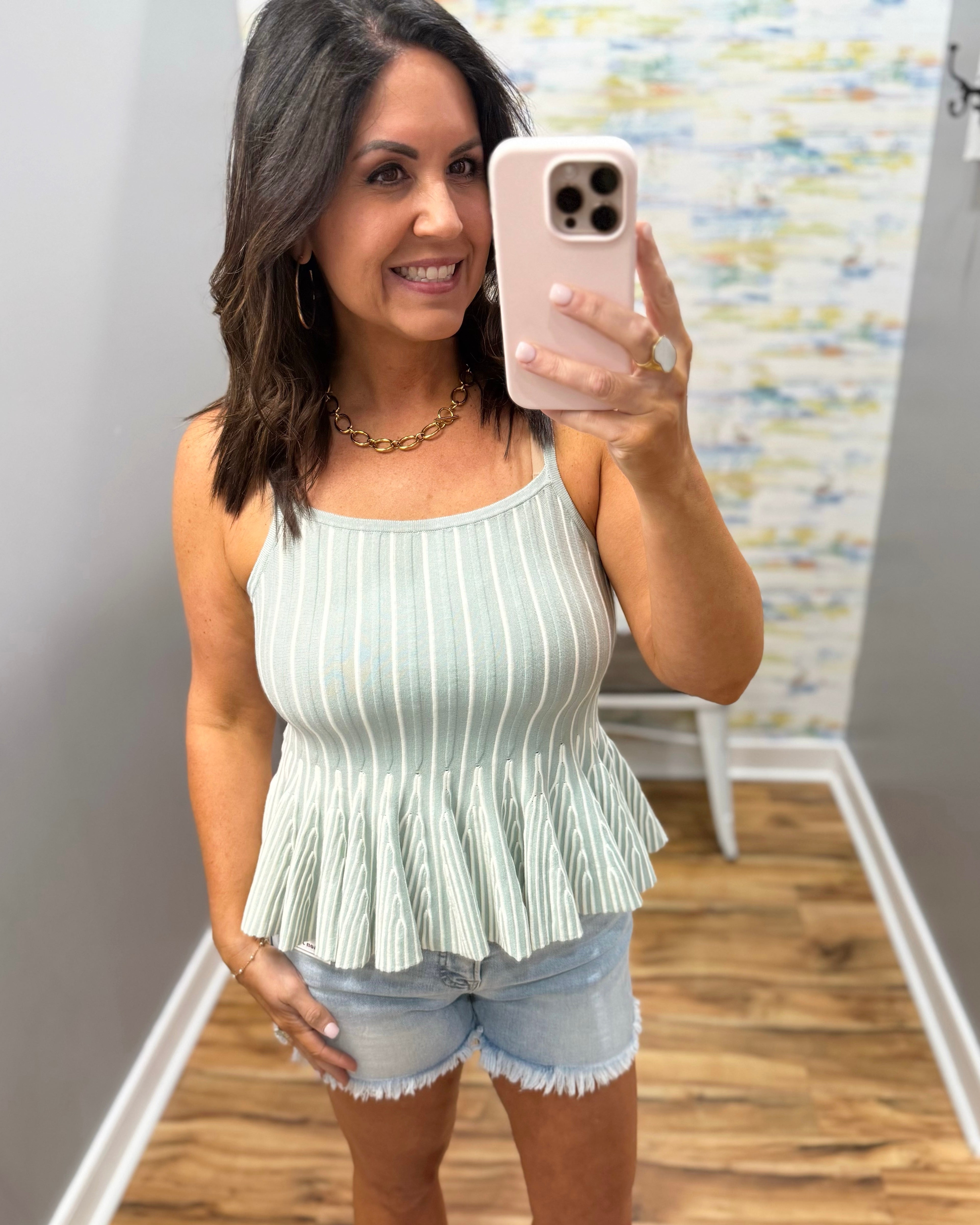 Woman taking a mirror selfie wearing a white pleated top and denim shorts.