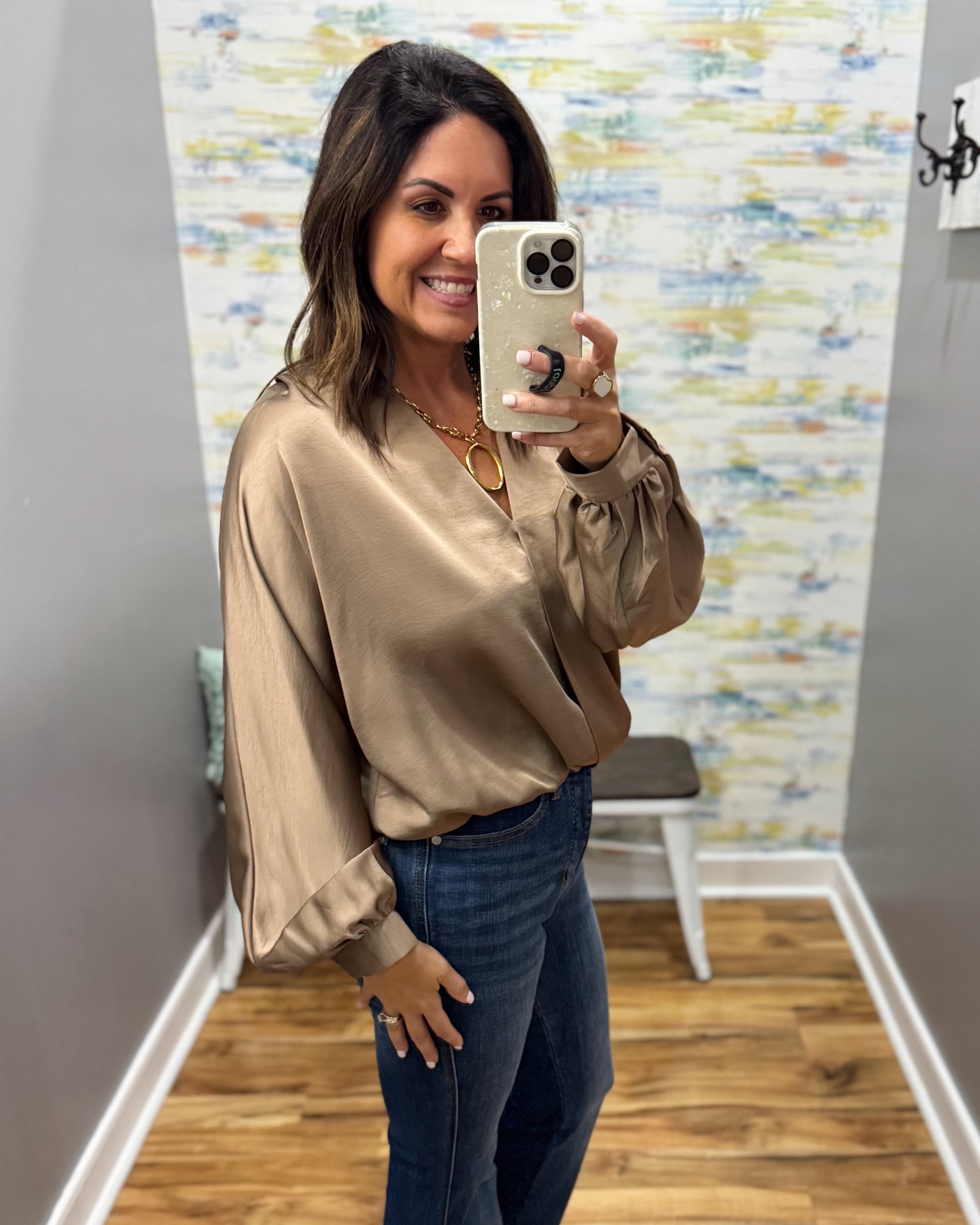 A woman takes a mirror selfie wearing a shiny taupe satin blouse and dark blue jeans in a home setting with a wood floor and abstract patterned wall.