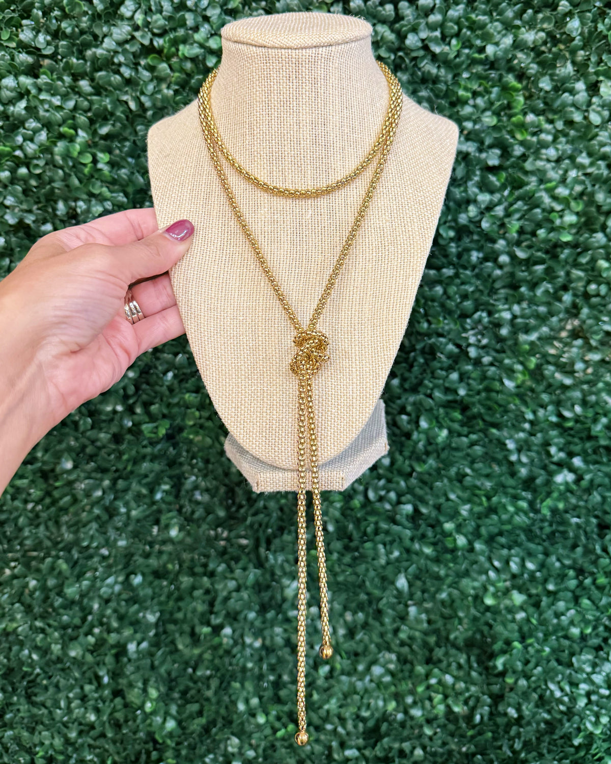 Chansuttpearls gold-toned double-strand necklace with a knotted center detail and long dangling chains, displayed on a beige fabric jewelry bust. A hand with pink nail polish is holding the left side of the bust, set against a green leaf wall background.