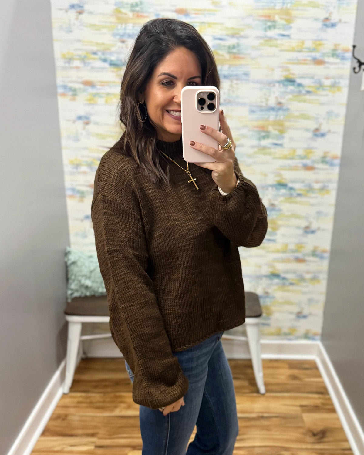 A woman takes a mirror selfie wearing a brown textured knit sweater and blue jeans in a home setting with a patterned wallpaper background.