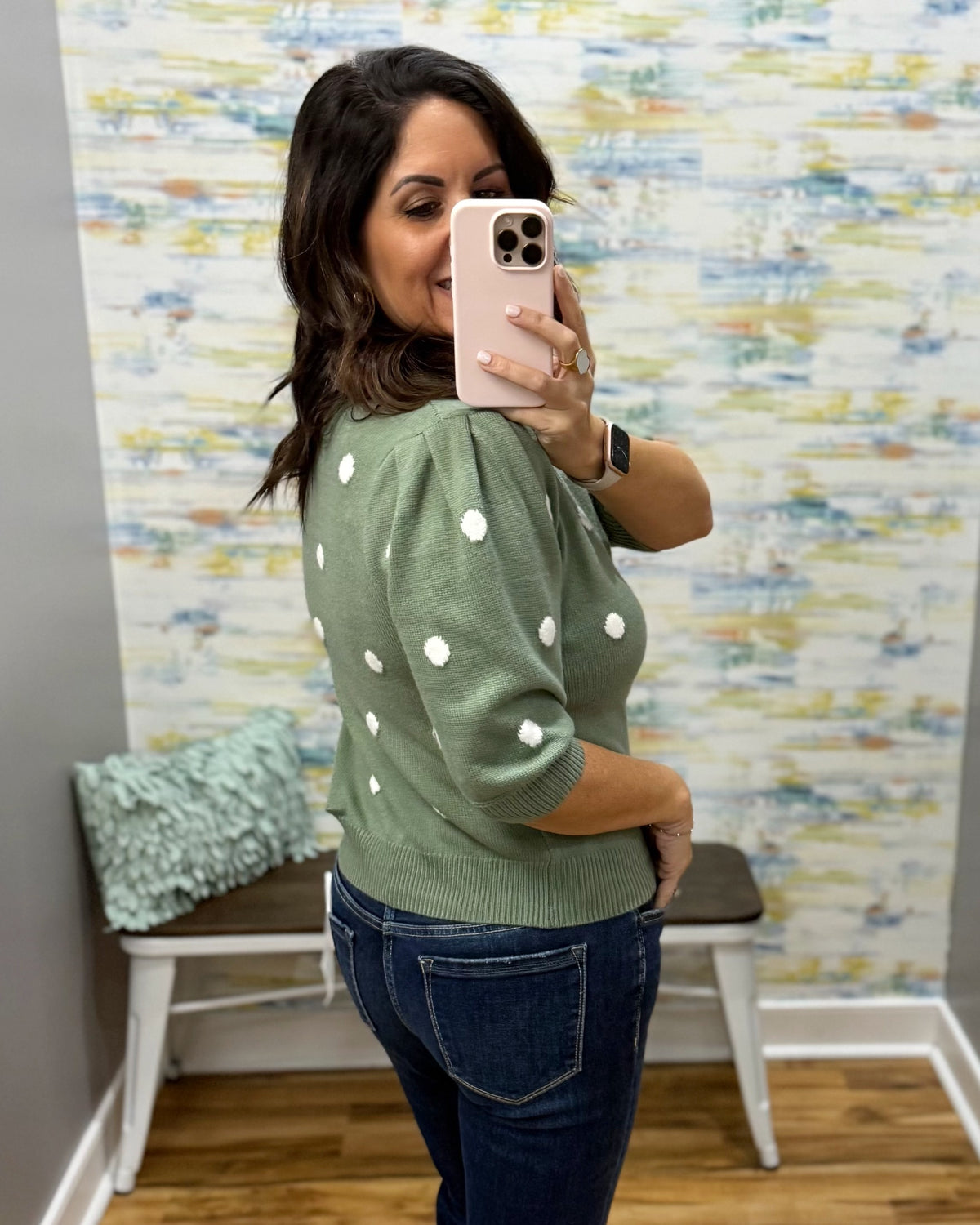 A woman takes a mirror selfie wearing a sage green knit sweater with white polka dots and dark blue jeans in a room with patterned wallpaper.