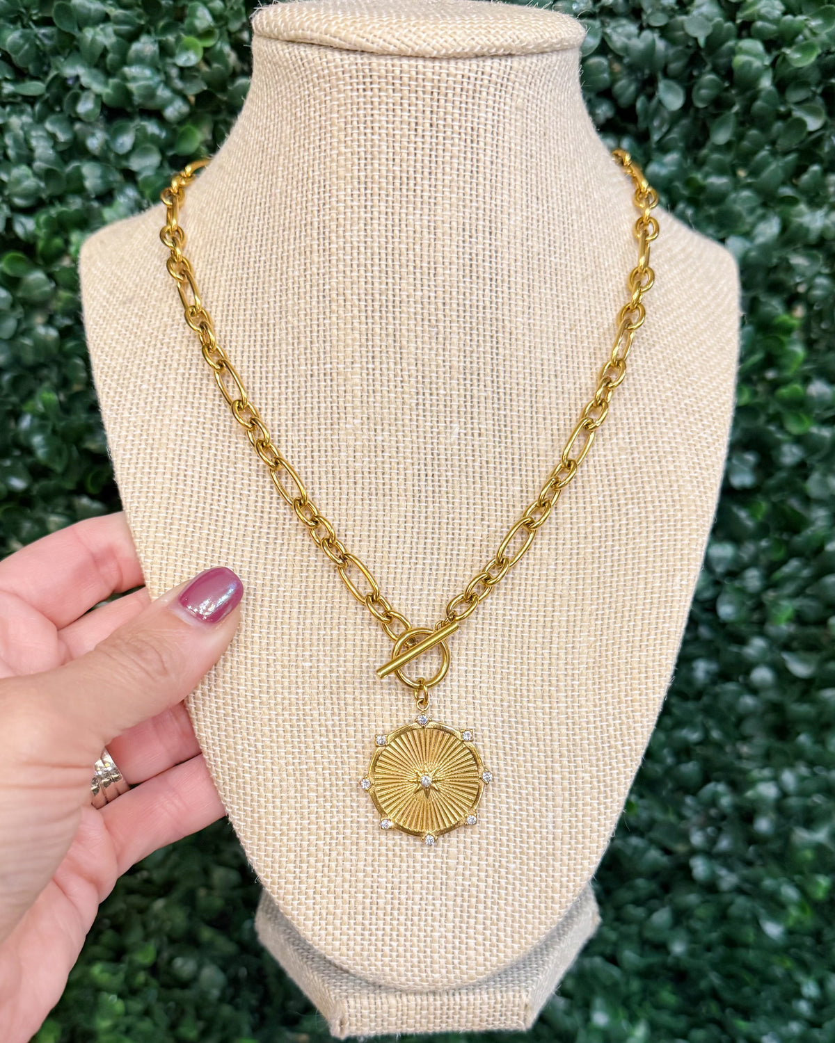 A gold chain necklace with a sunburst pendant and toggle clasp, displayed on a beige fabric jewelry bust. A hand with purple nail polish is holding the left side of the bust, and a green leafy wall serves as the background.
