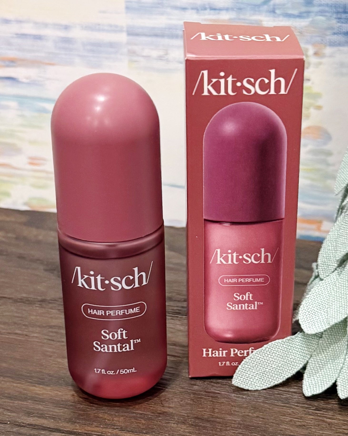 A bottle of Kit-sch Soft Santal hair perfume next to its retail box, both in a dusty rose pink color, displayed on a wooden surface with a decorative leaf in the foreground.