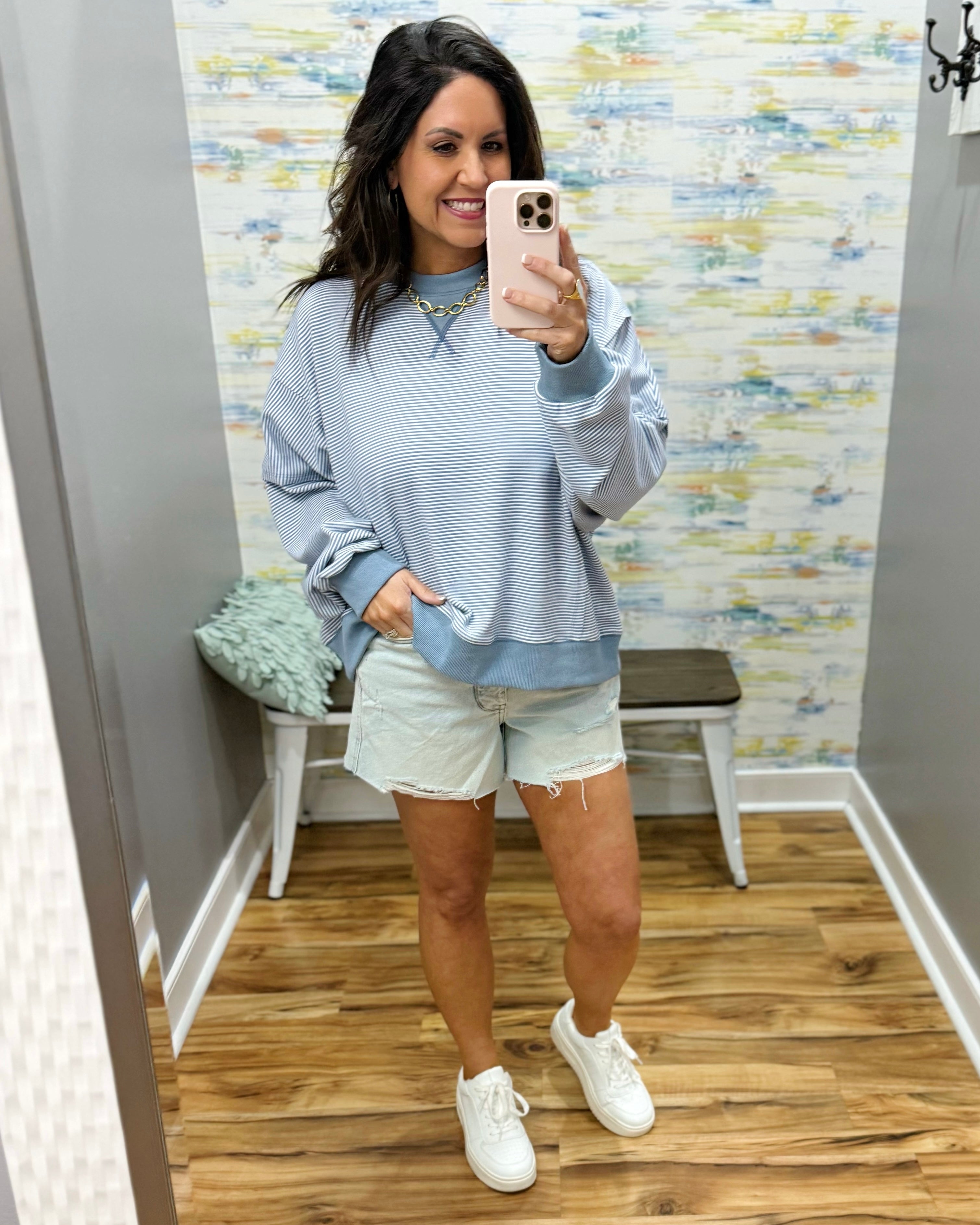 Woman taking a mirror selfie wearing a blue sweater, light shorts, and white sneakers in a room with patterned wallpaper.