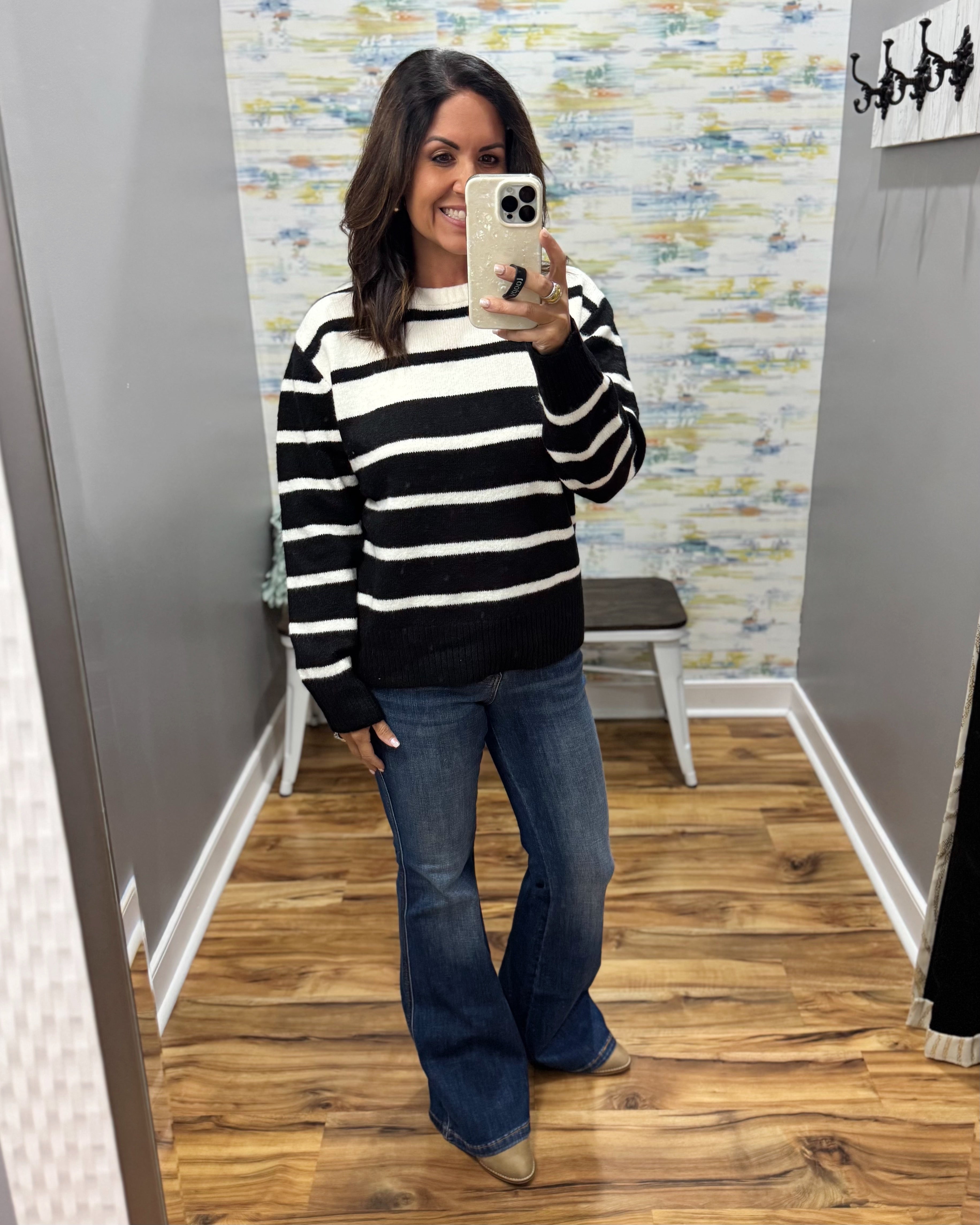 A woman takes a mirror selfie wearing a black and white horizontally striped knit sweater, blue flare jeans, and tan boots in a fitting room with wood floors and abstract wallpaper.