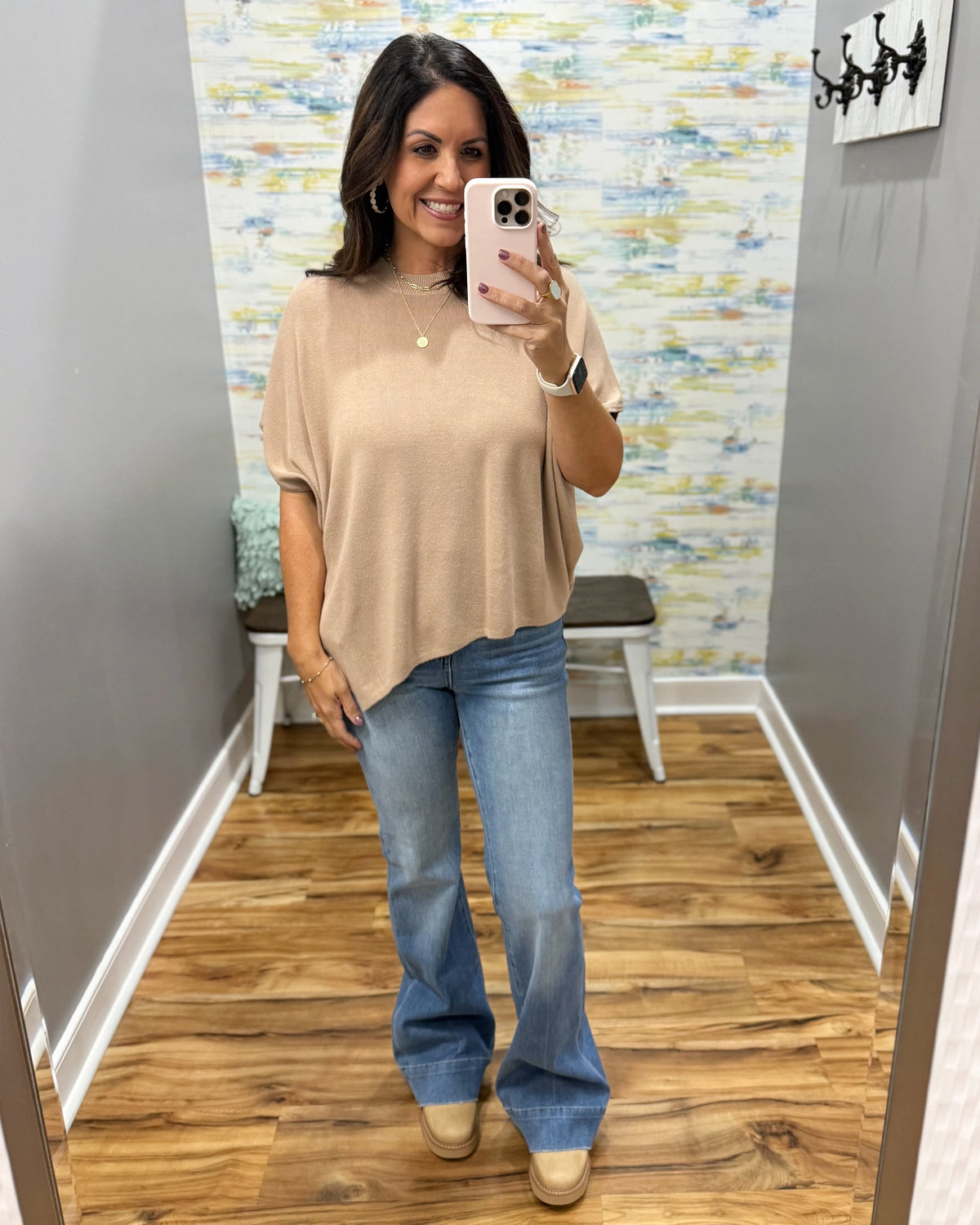 A woman takes a mirror selfie in a fitting room wearing a beige short-sleeve knit top and blue flare jeans.