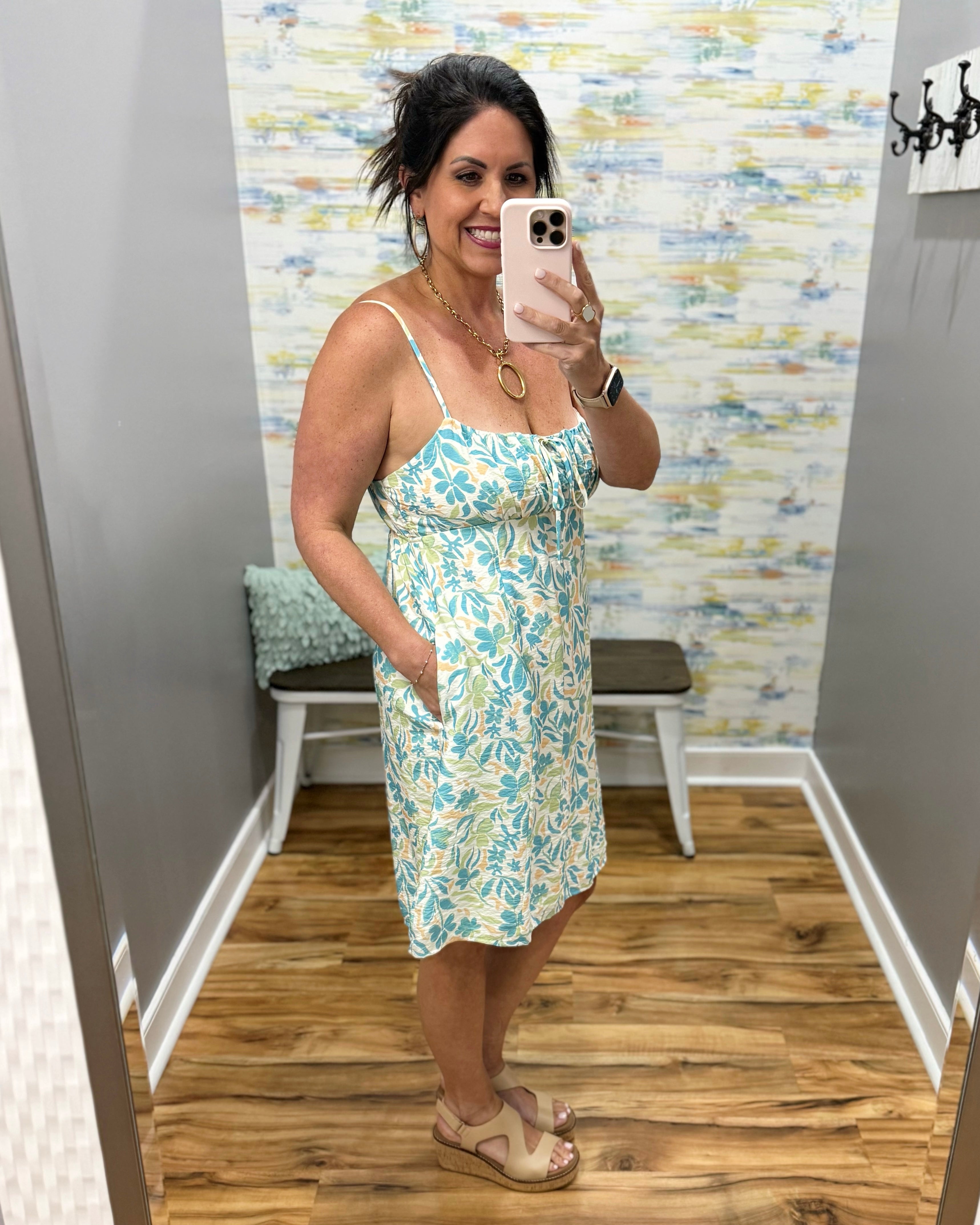 Woman taking a mirror selfie wearing a floral dress in a room with a textured wall.