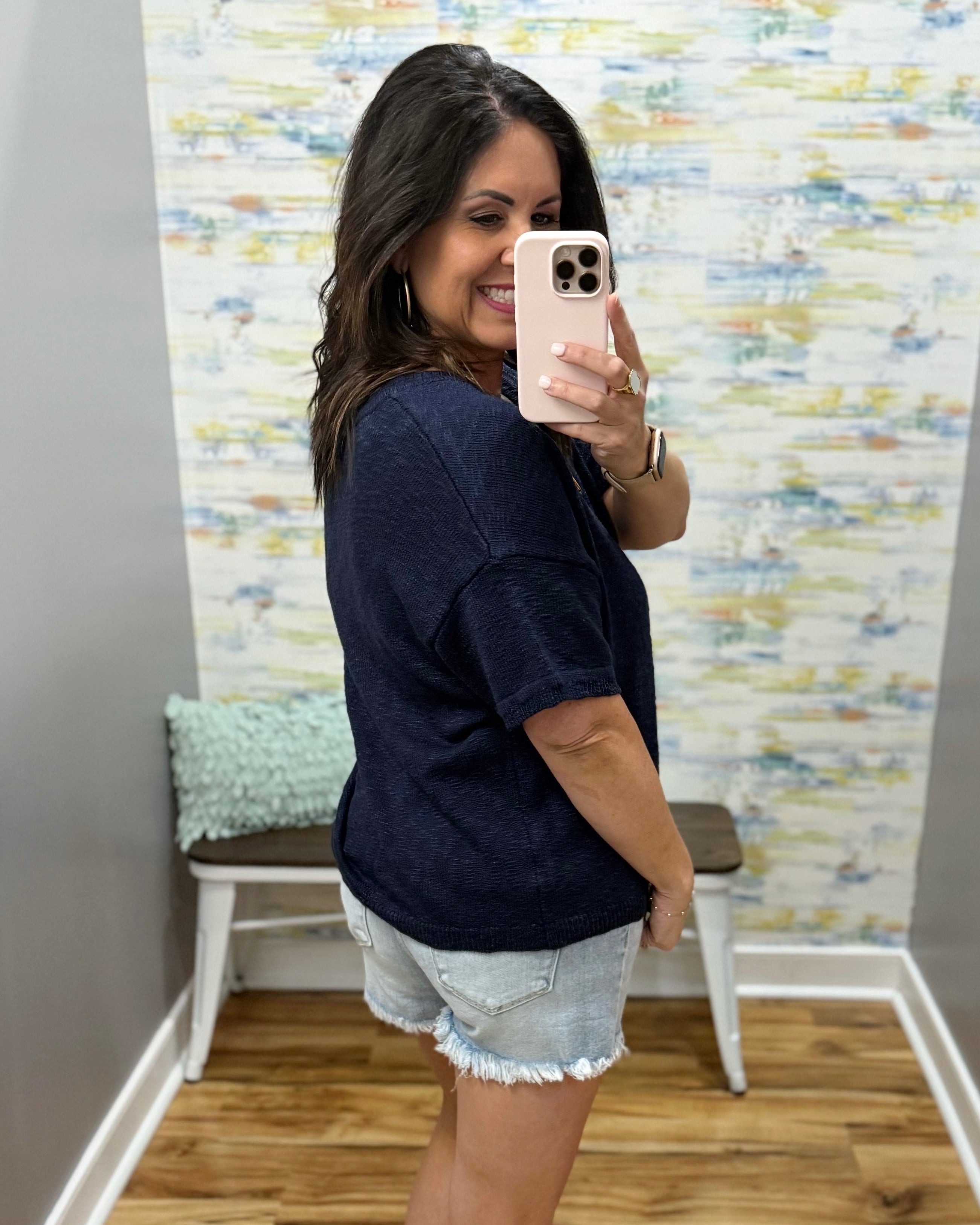 Woman taking a mirror selfie wearing a navy blue shirt and denim shorts in a room with a textured wall.