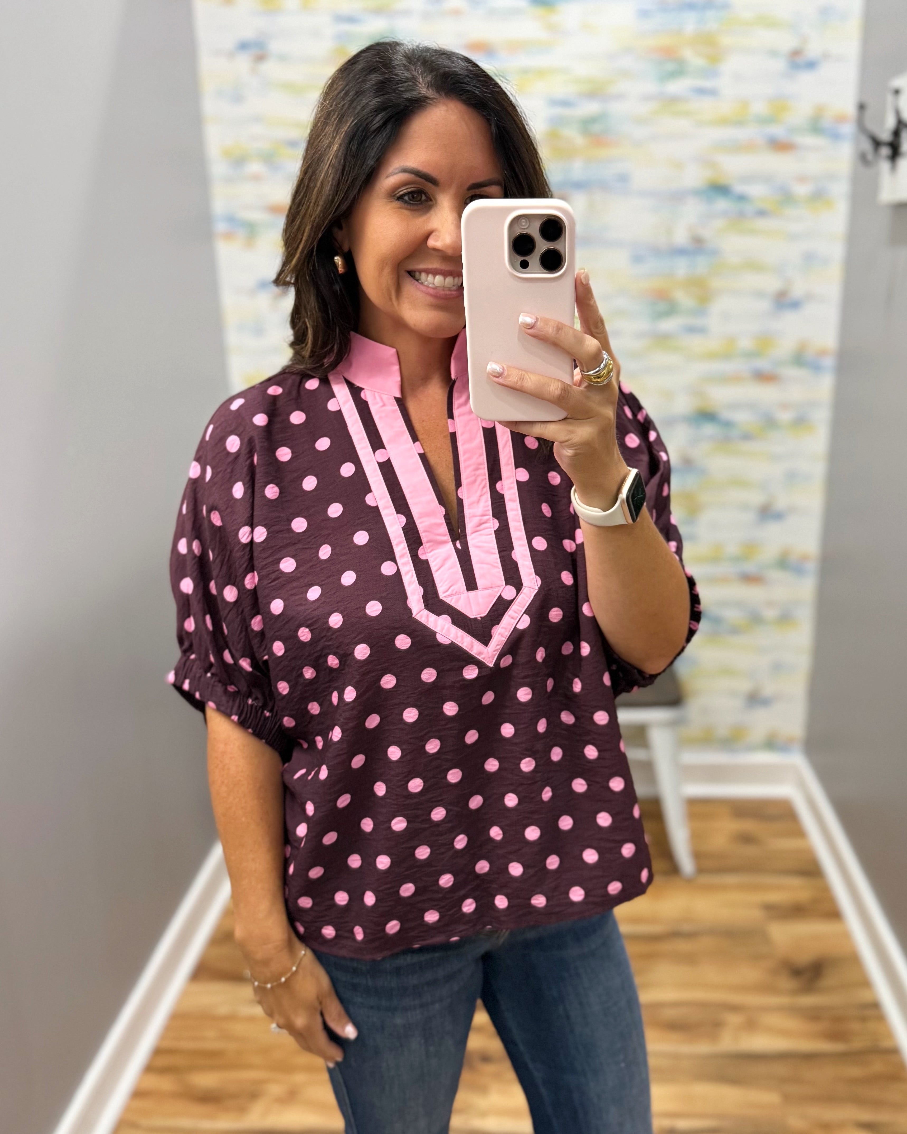 A woman takes a mirror selfie wearing a short-sleeve blouse with pink polka dots and a pink V-neckline detail on a dark plum background, paired with blue jeans.