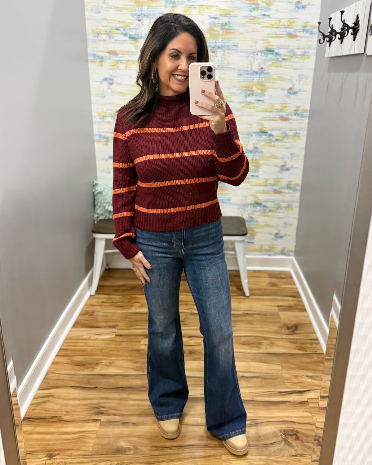 A woman takes a mirror selfie wearing a burgundy and orange striped mock neck sweater, blue flare jeans, and beige boots in a fitting room with wood floors and a colorful abstract wall.