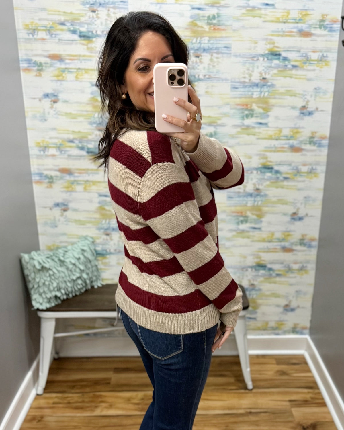 A woman takes a mirror selfie wearing a beige and burgundy striped knit sweater, blue jeans, and a light pink phone case in front of an abstract patterned wall.