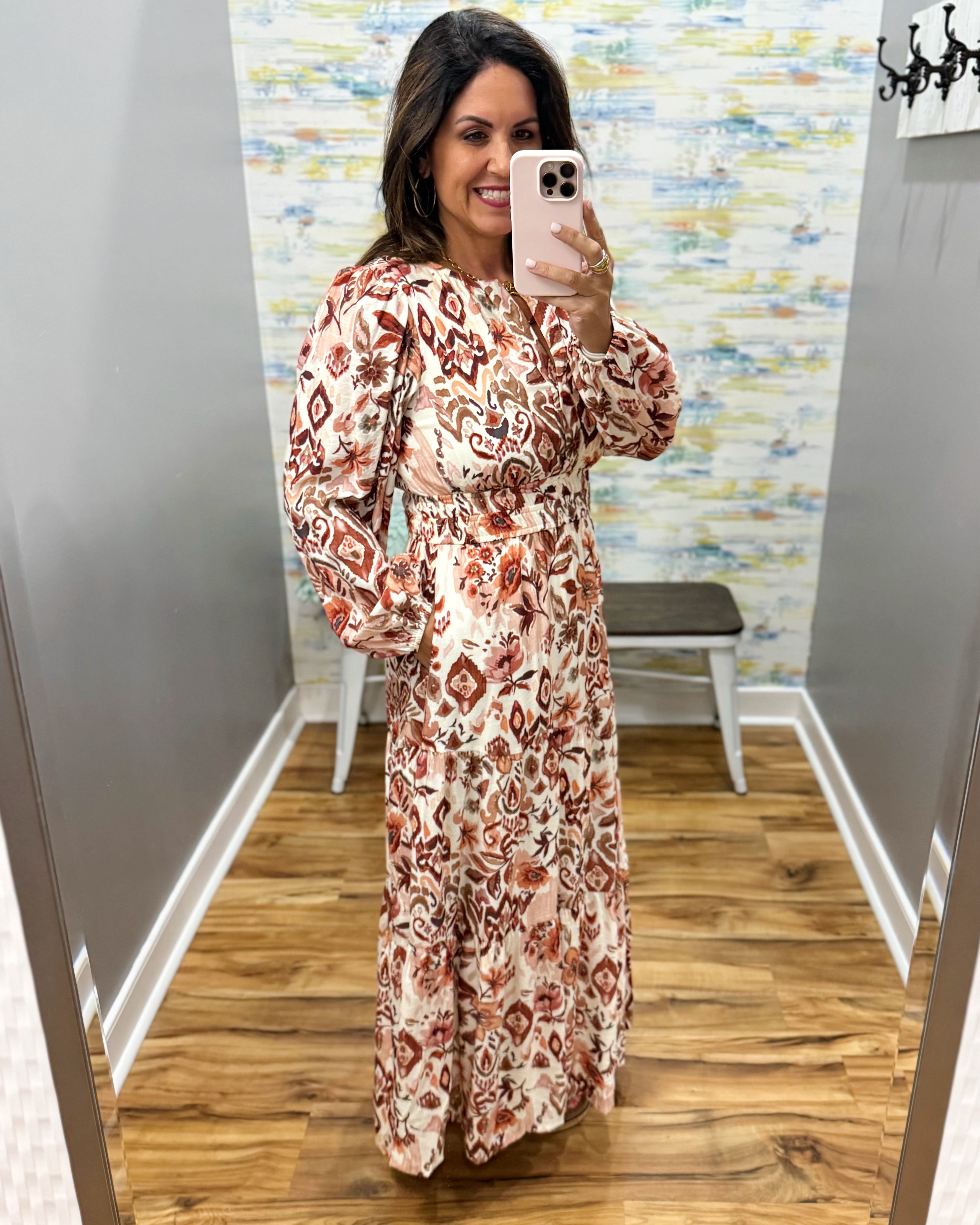 A woman takes a mirror selfie in a fitting room wearing a long-sleeve maxi dress with a rust red and beige geometric pattern on a cream background.