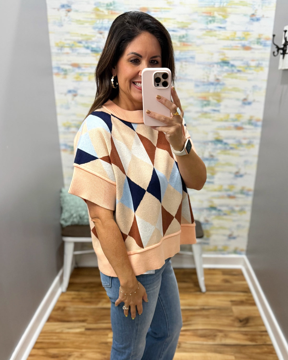 A woman takes a mirror selfie wearing a short-sleeve knit sweater with a geometric diamond pattern in peach, beige, navy, and brown, paired with blue jeans.