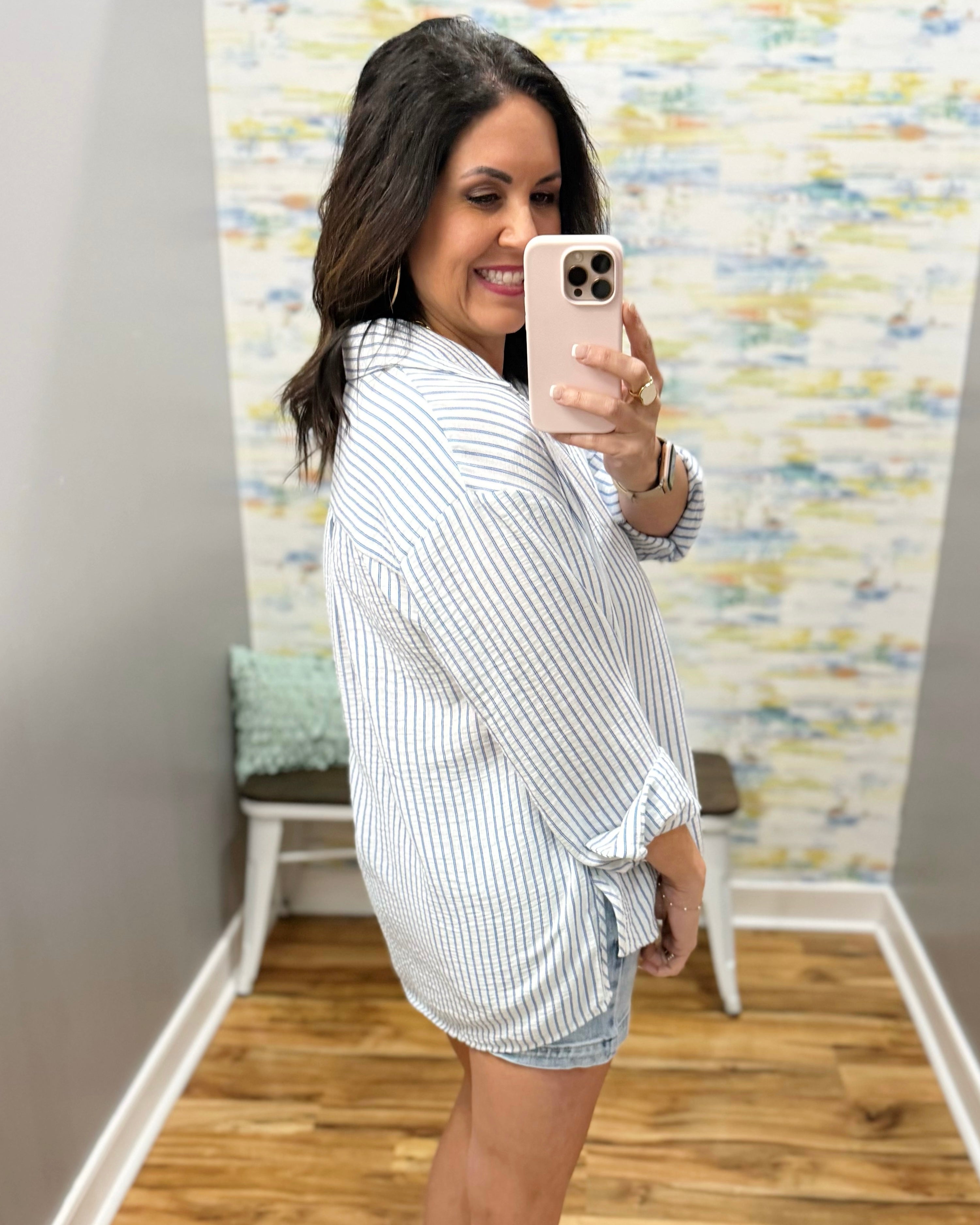 Woman taking a mirror selfie wearing a striped shirt and shorts in a room with a colorful wall.