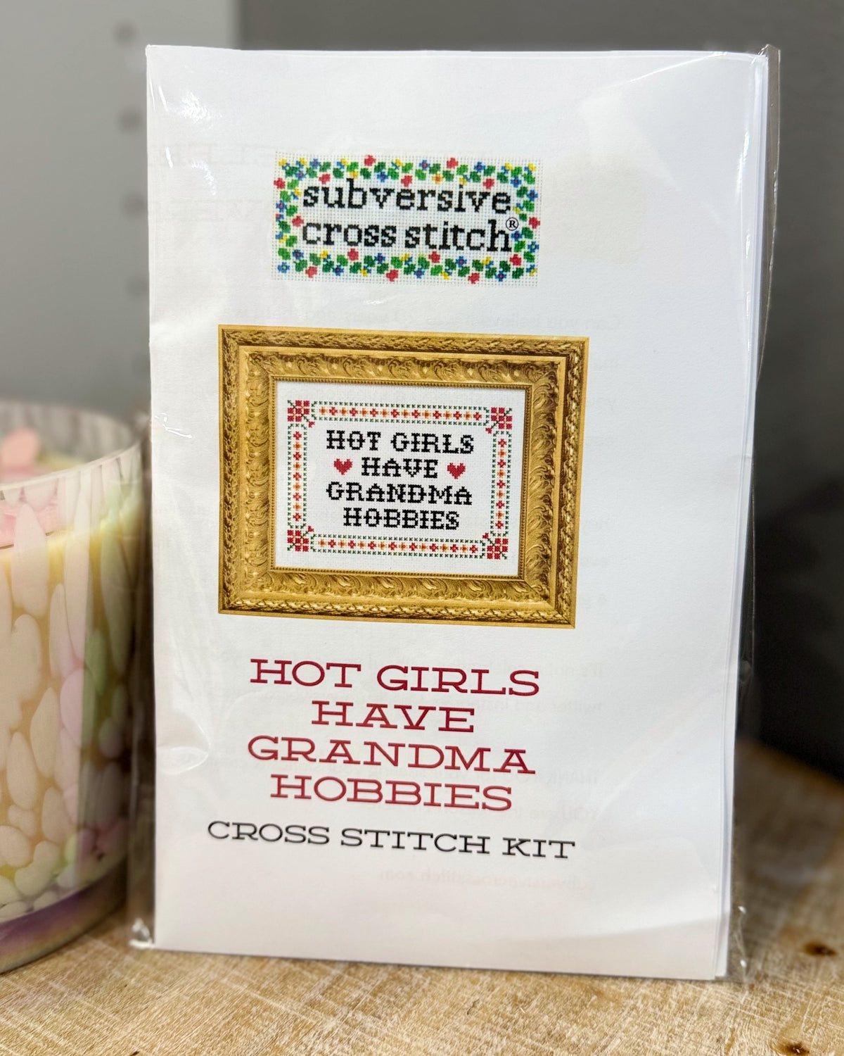 A packaged cross stitch kit in a clear plastic sleeve. The front shows a white card with a framed sample cross stitch design reading 'HOT GIRLS HAVE GRANDMA HOBBIES' and the brand name 'subversive cross stitch' at the top.