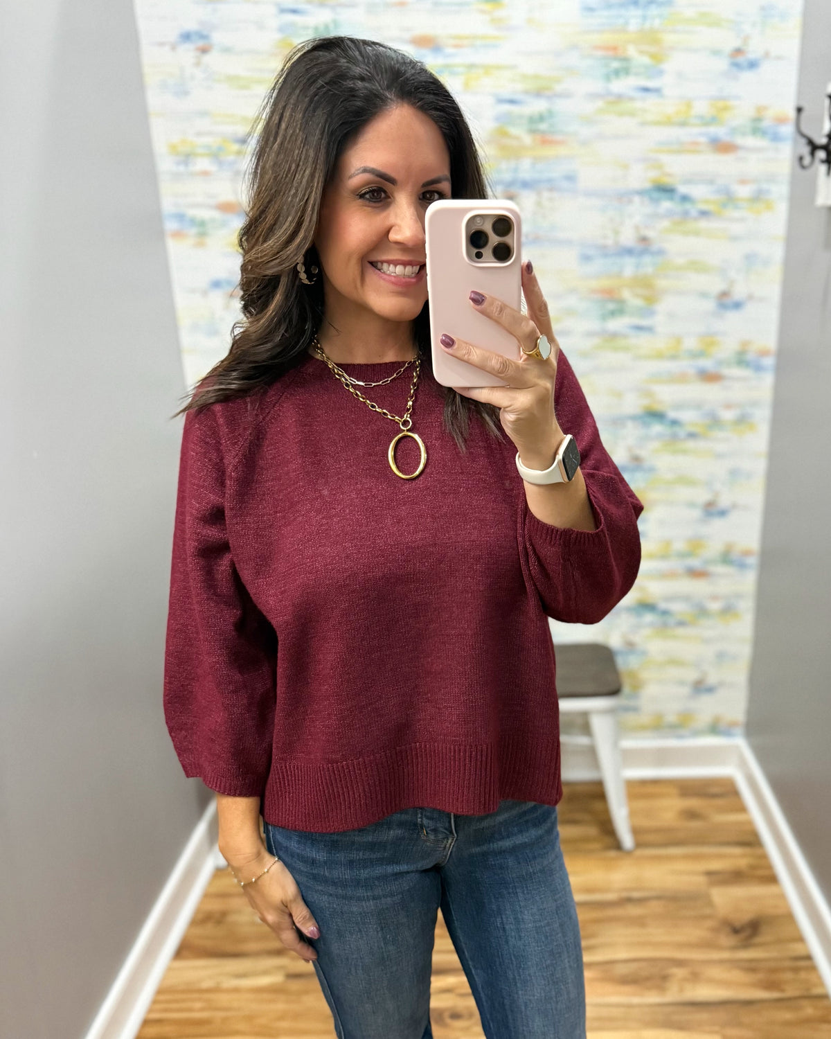 A woman takes a mirror selfie wearing a heathered burgundy knit sweater, blue jeans, and gold jewelry in a home setting with a colorful abstract wallpaper.
