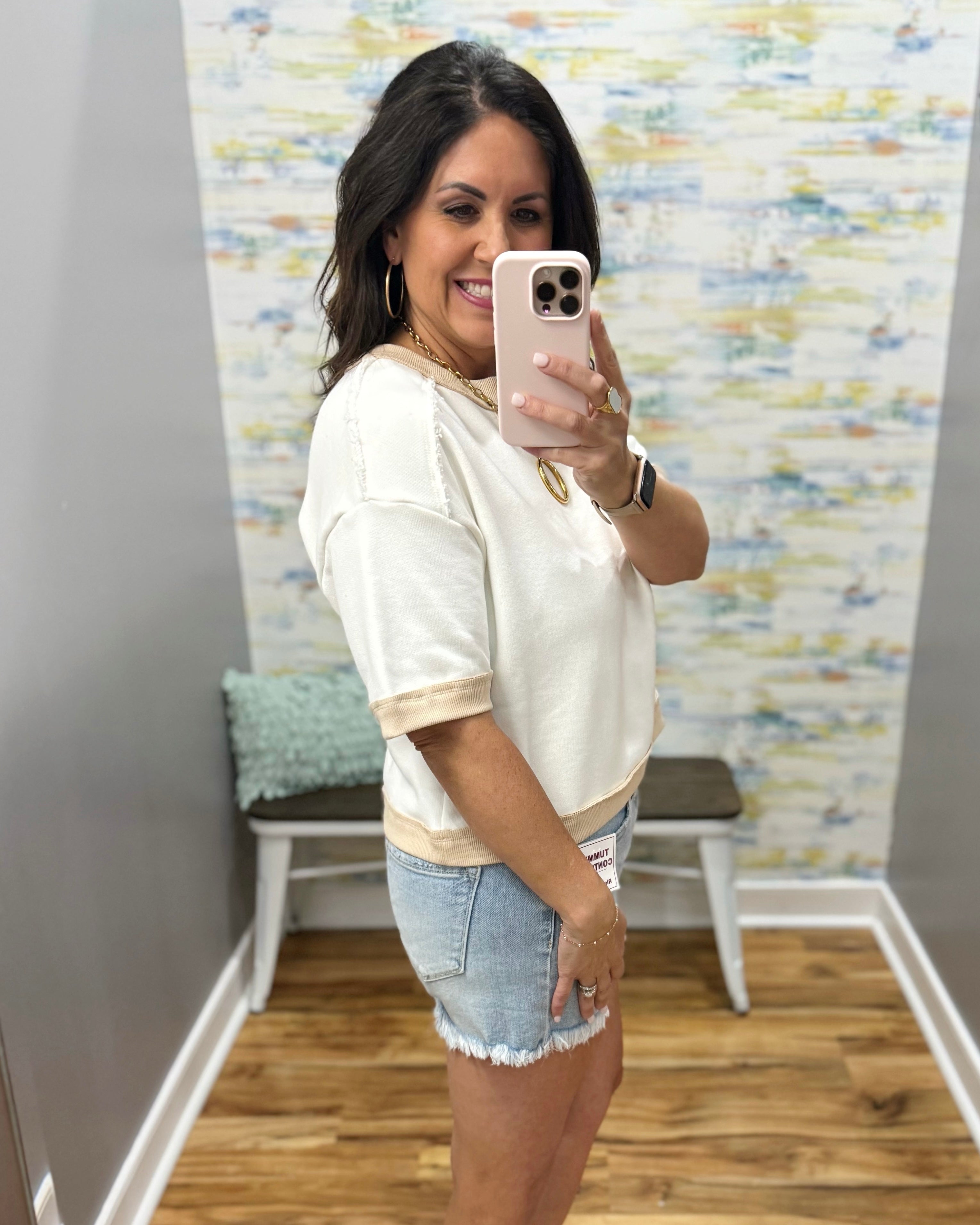 Woman taking a mirror selfie wearing a white top and denim shorts in a room with a colorful wall.