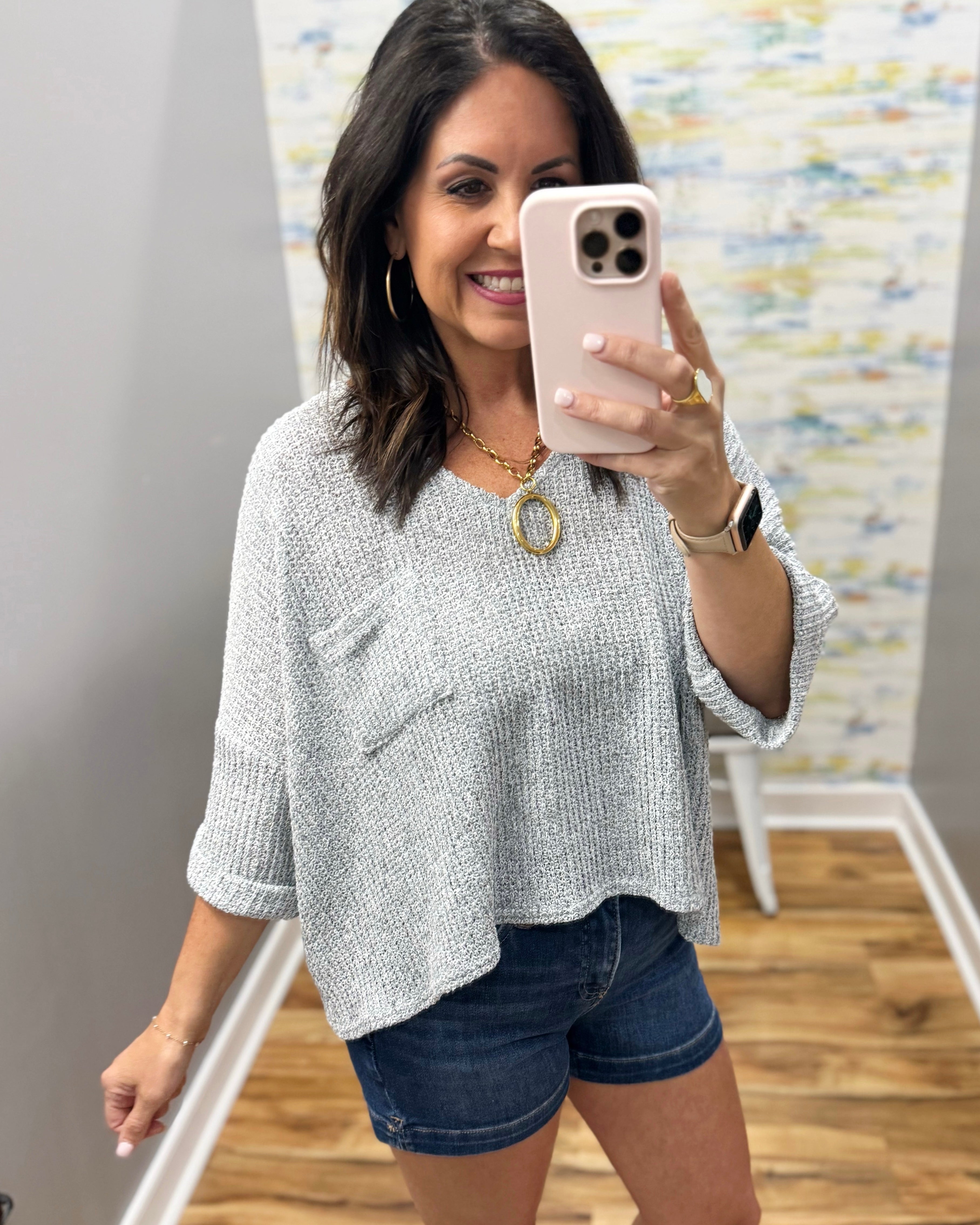 Woman taking a mirror selfie wearing a gray sweater and denim shorts.