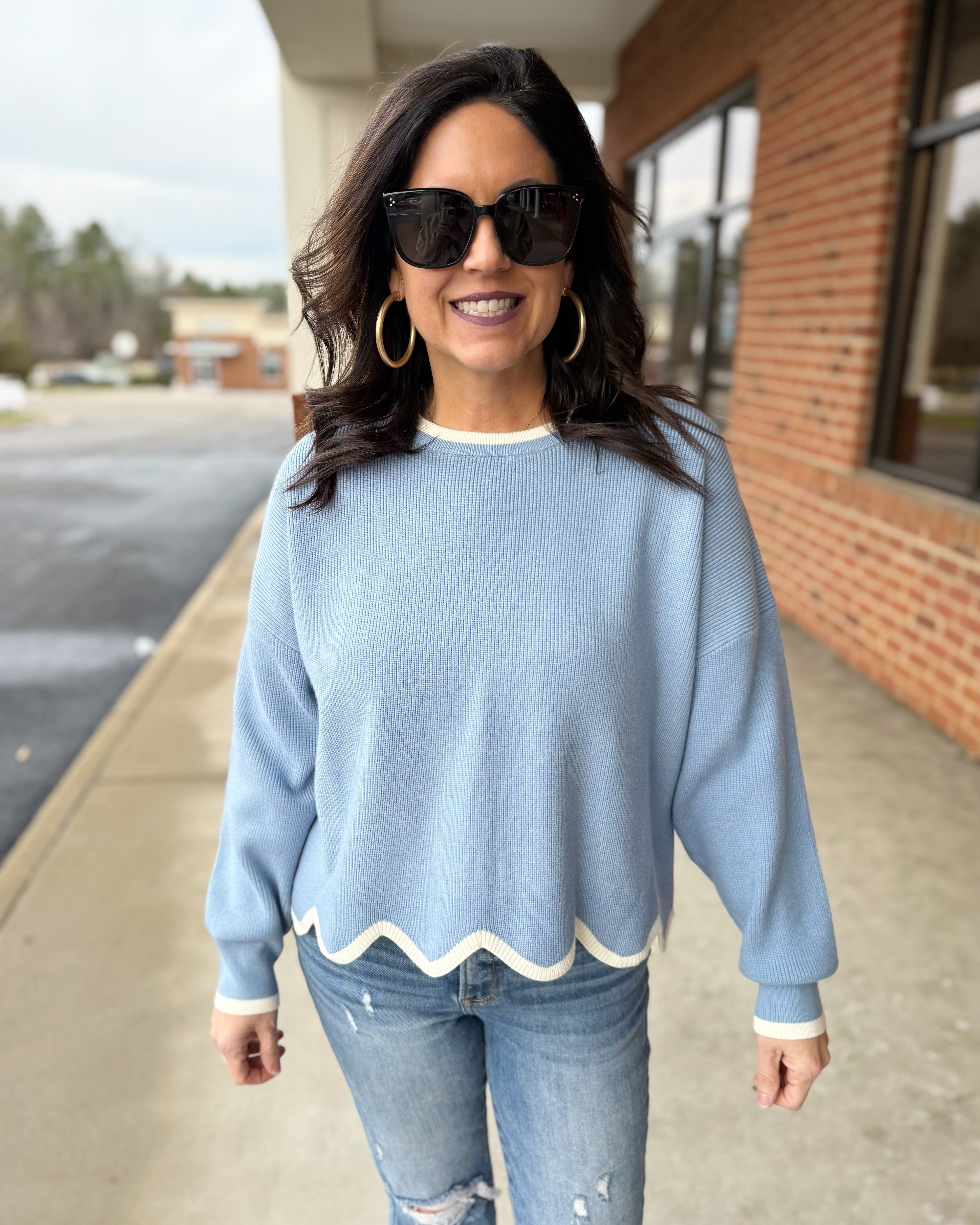 Woman wearing a light blue sweater with scalloped hem and sunglasses outdoors.