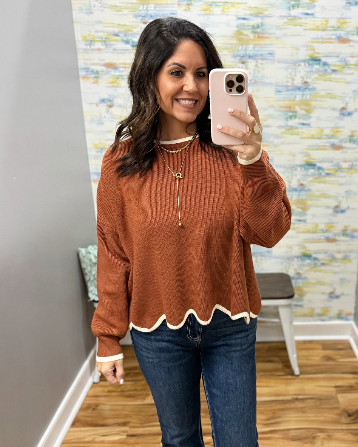 A woman takes a mirror selfie wearing a rust-orange knit sweater with a scalloped white hem and cuffs, paired with blue jeans and gold necklaces.
