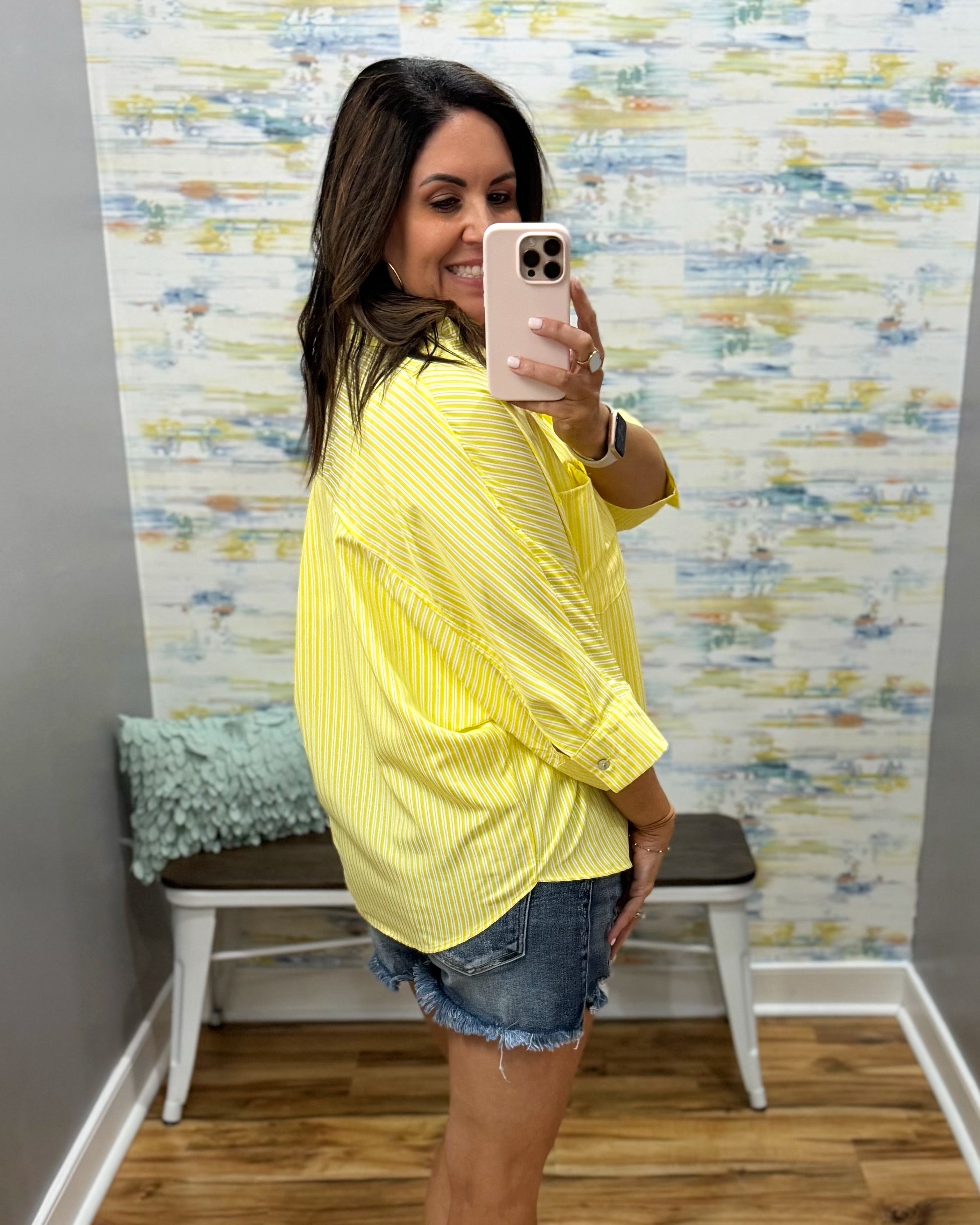 Woman taking a mirror selfie wearing a yellow sweater and denim shorts in a room with colorful wall art.