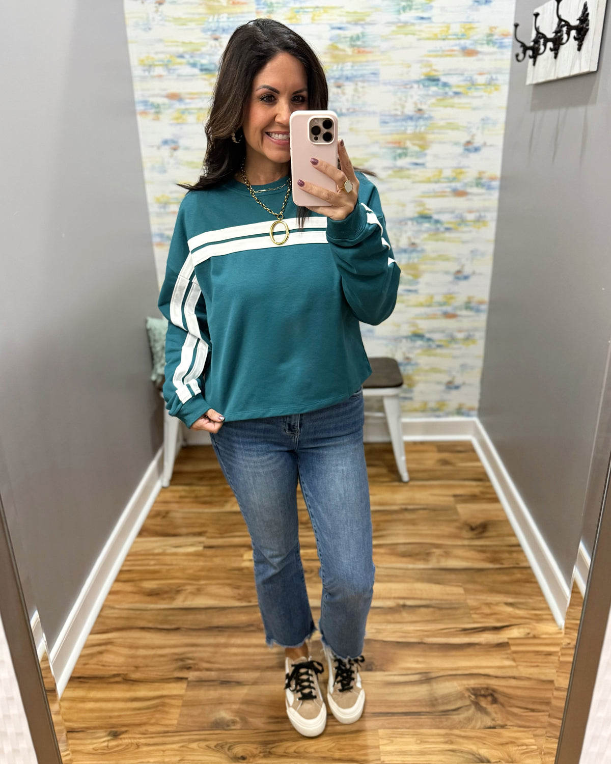 A woman takes a mirror selfie wearing a teal long-sleeve sweatshirt with white athletic stripes on the sleeves, paired with blue jeans and white sneakers.