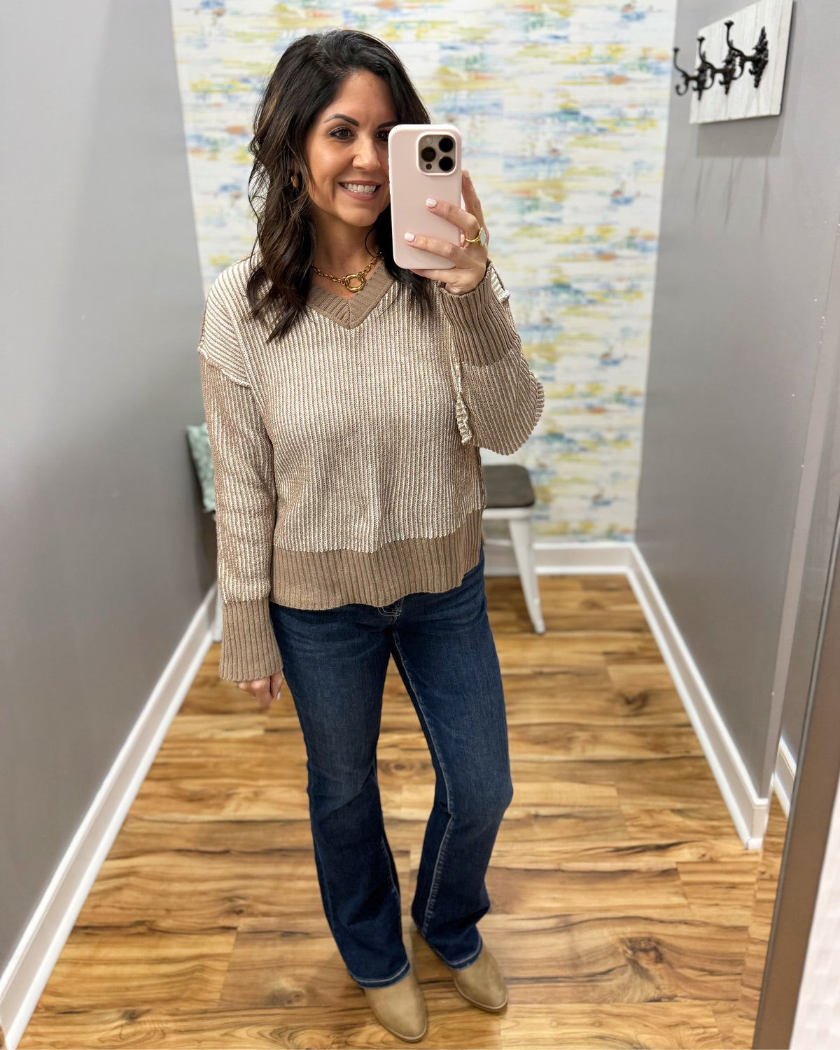 A woman takes a mirror selfie in a home hallway wearing a beige ribbed knit sweater, dark blue jeans, and tan ankle boots.