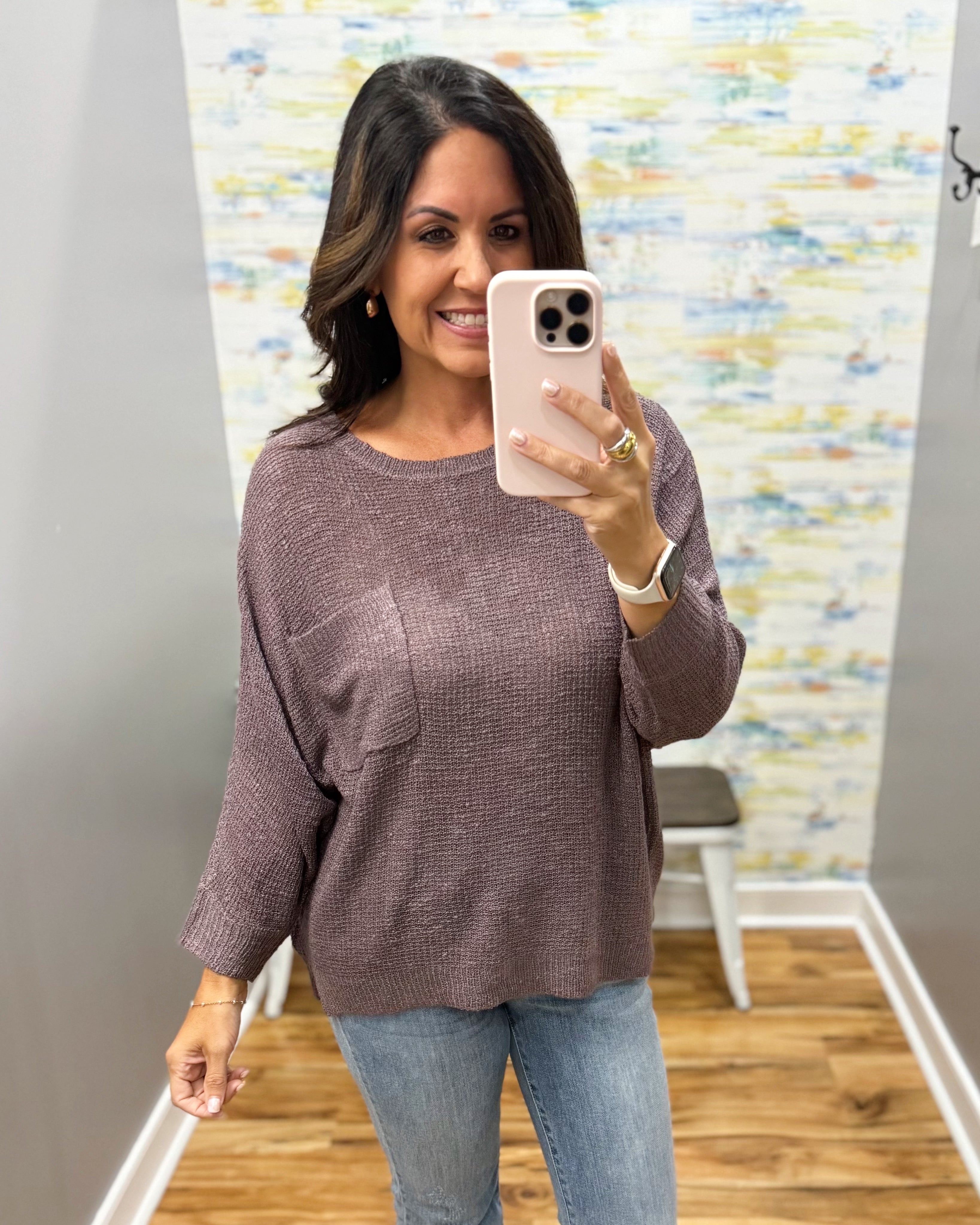 A woman takes a mirror selfie wearing a textured, long-sleeve crewneck top in a dusty mauve-brown color, paired with blue jeans. The background shows a gray wall, a colorful abstract print, and a wooden floor.