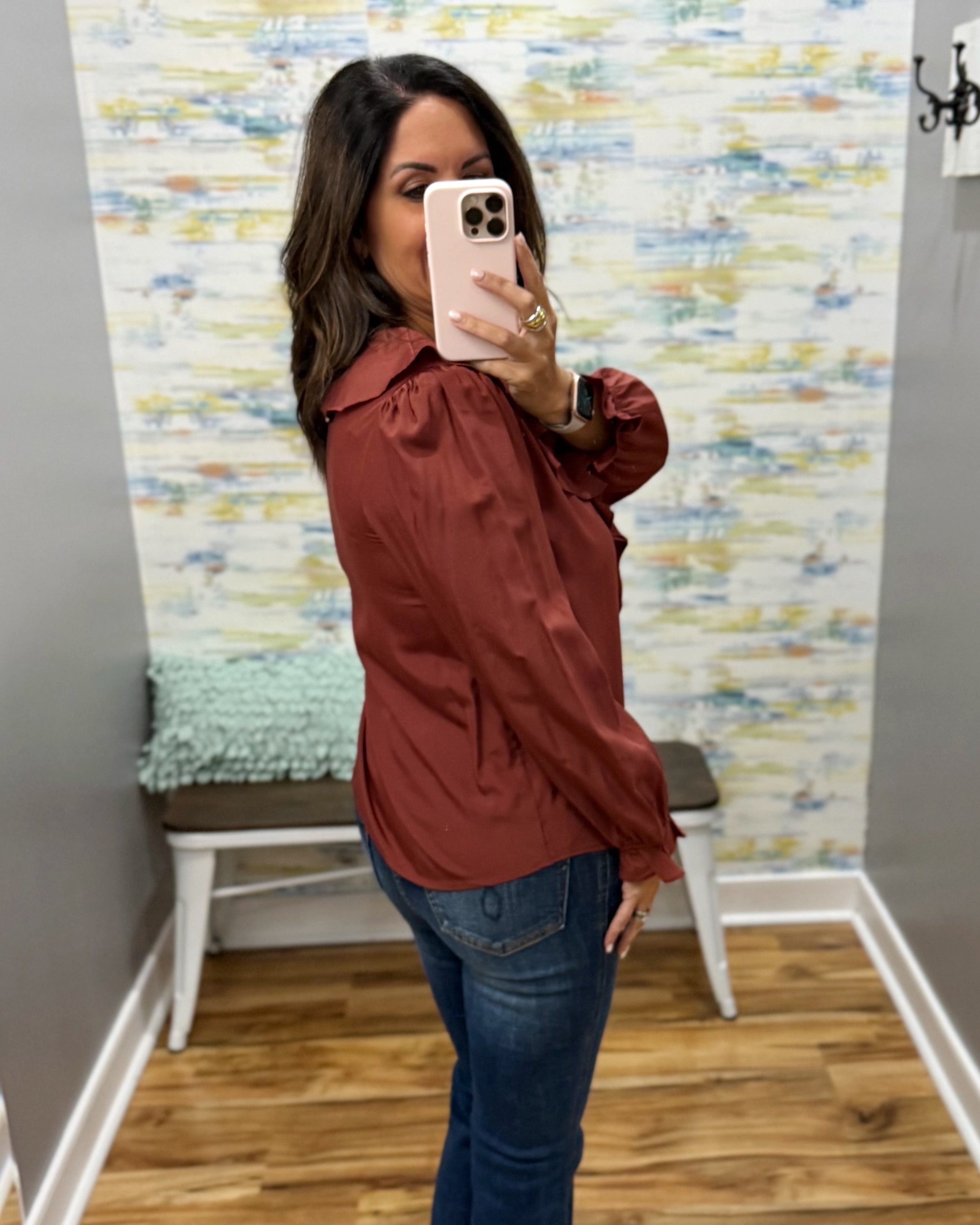 A woman takes a mirror selfie in a fitting room wearing a rust-colored long-sleeve blouse and blue jeans.
