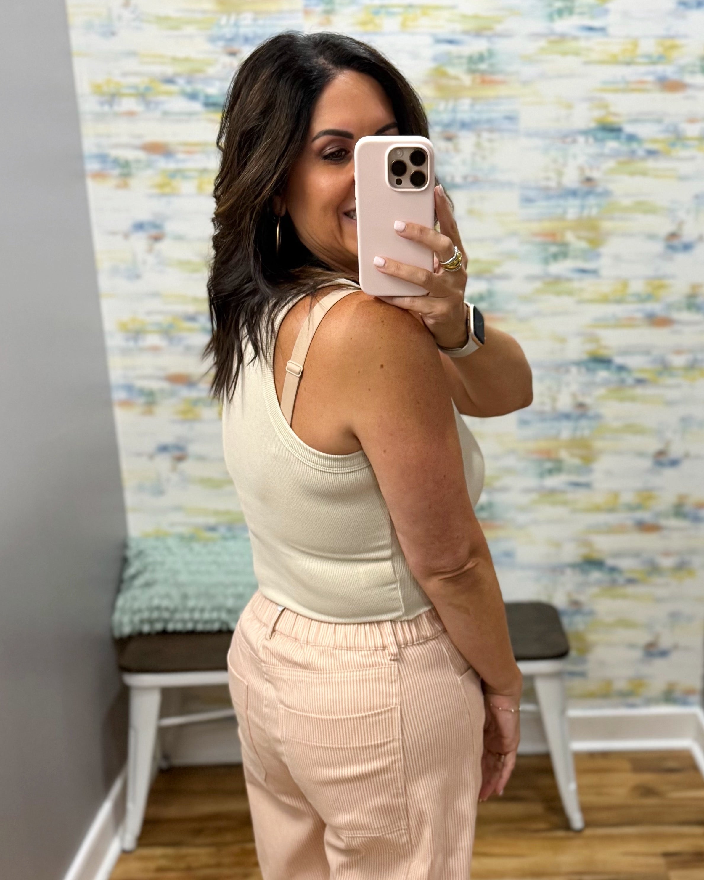 A woman taking a mirror selfie in a fitting room, wearing a cream ribbed tank top and light pink corduroy pants. The background features a patterned wallpaper and a small bench.