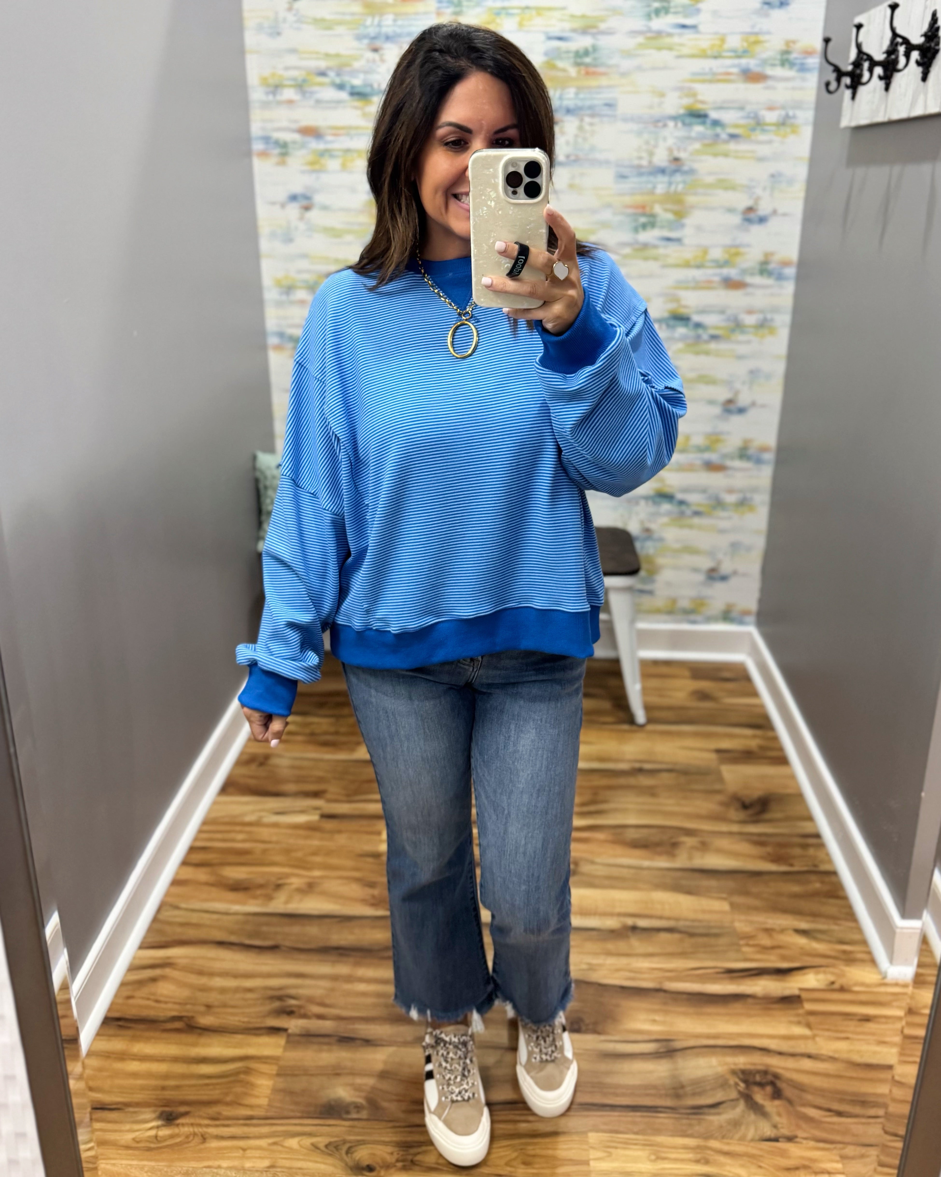 A woman takes a mirror selfie wearing a bright blue crewneck sweatshirt, blue jeans, and beige sneakers in a home hallway.