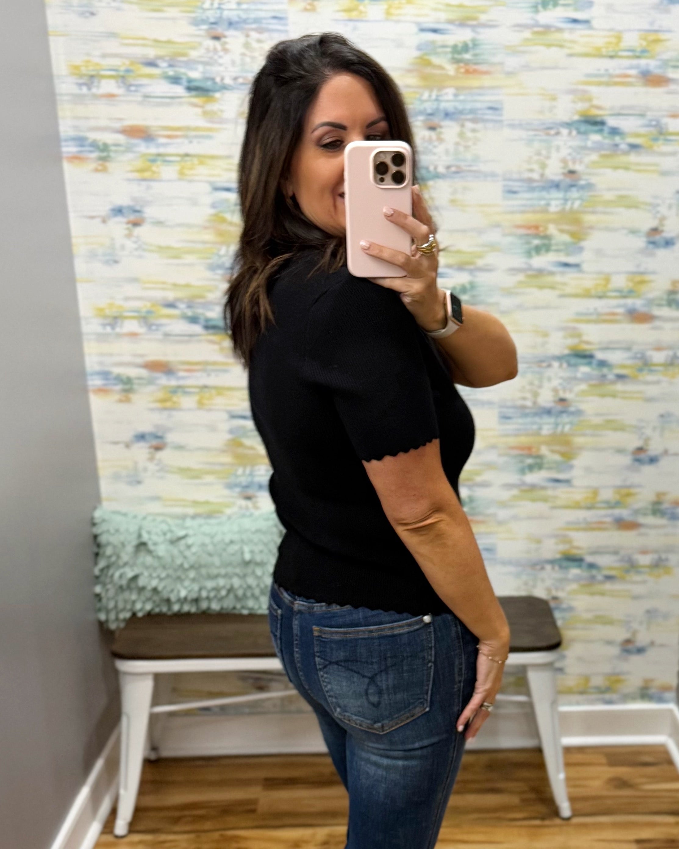 A woman takes a mirror selfie wearing a black short-sleeve knit top with a scalloped hem and blue jeans, standing in front of a colorful abstract wallpapered wall.