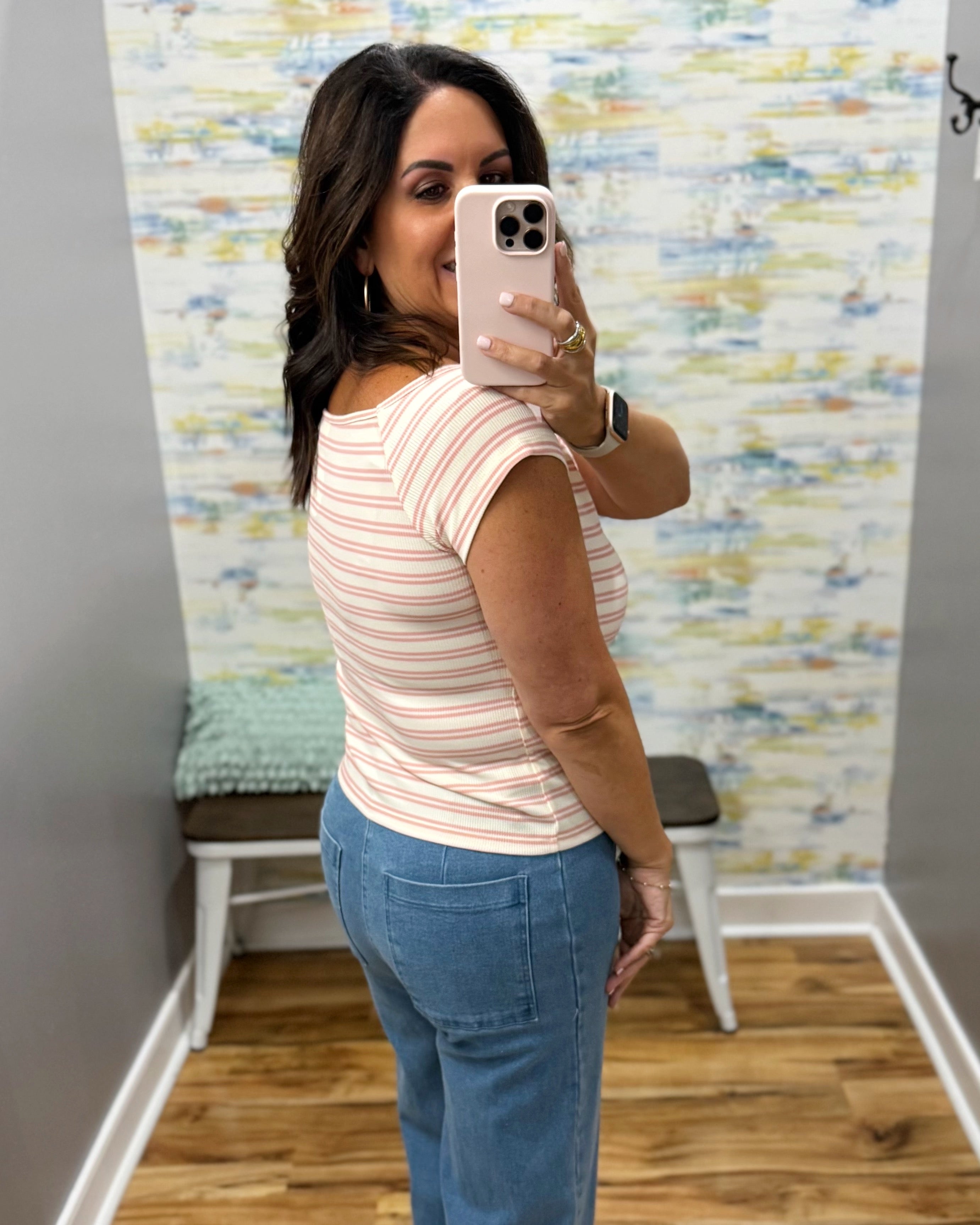 A woman taking a mirror selfie in a fitting room wearing a short-sleeve crewneck top with thin pink and white horizontal stripes, paired with blue jeans.