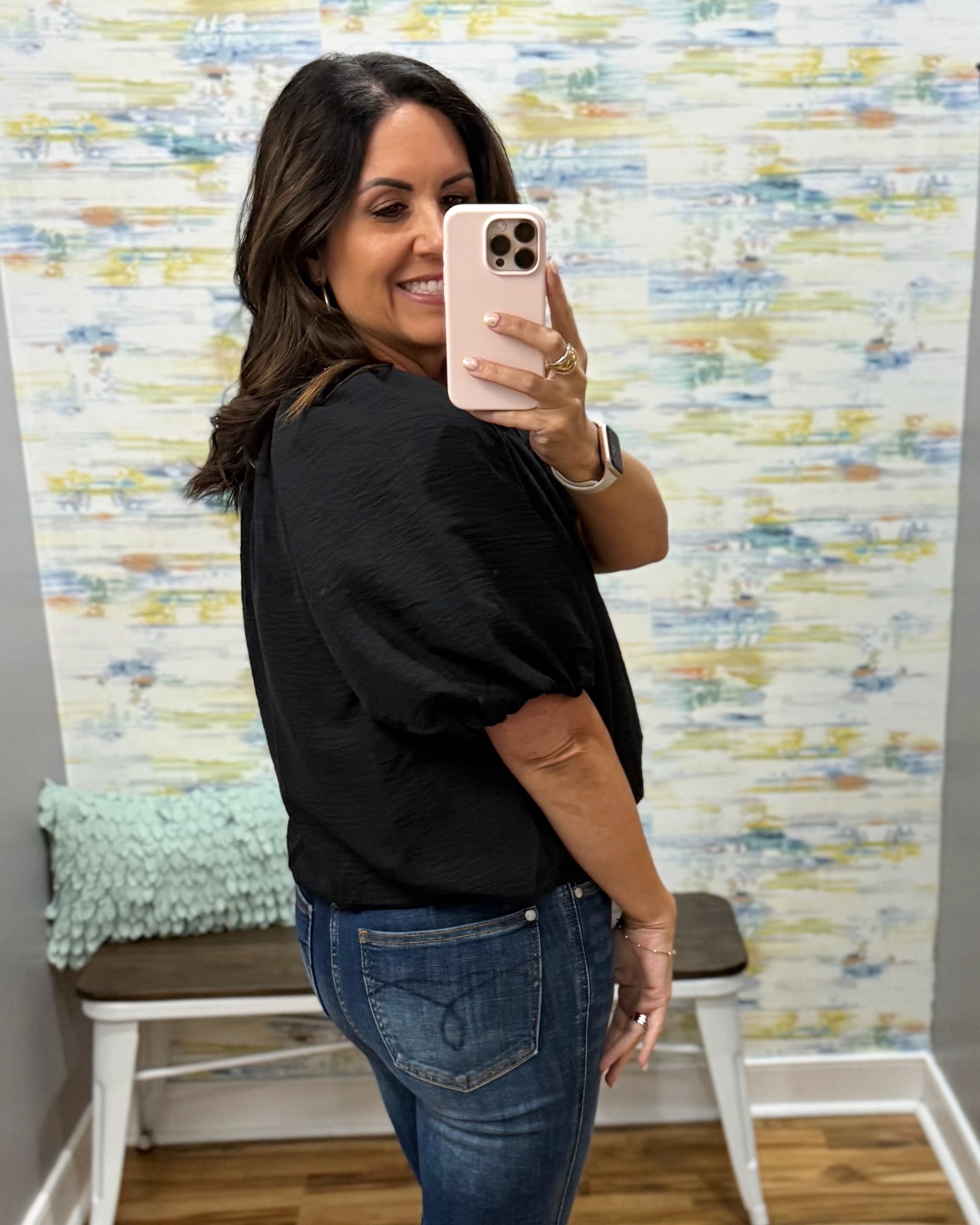 A woman taking a mirror selfie in a fitting room, wearing a black textured short-sleeve top and blue jeans.